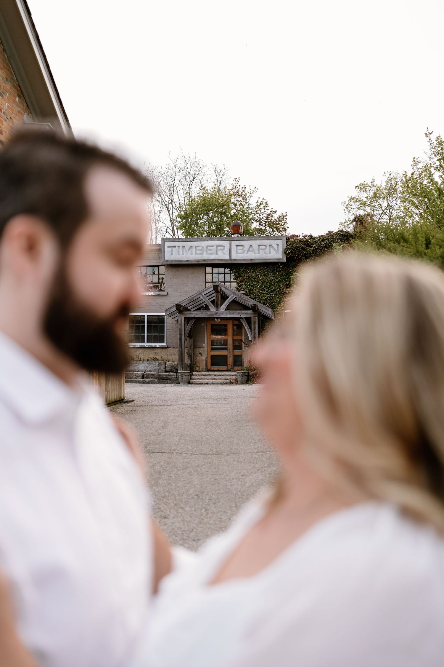 Couple in white shares a tender moment with the rustic Timber Barn venue blurred in the background.