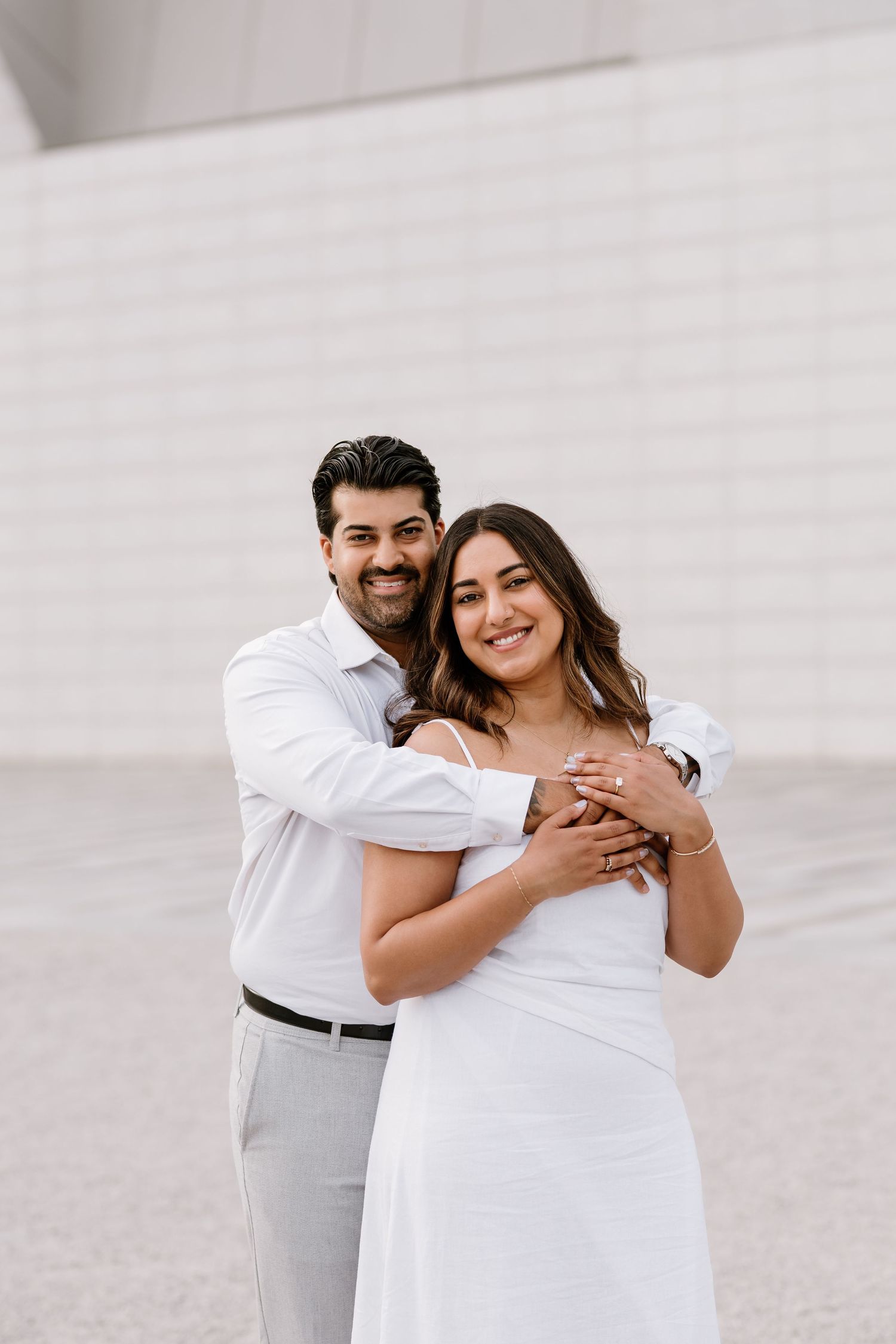 Couple in white outfits embracing outdoors, man hugging woman from behind, both smiling at camera.