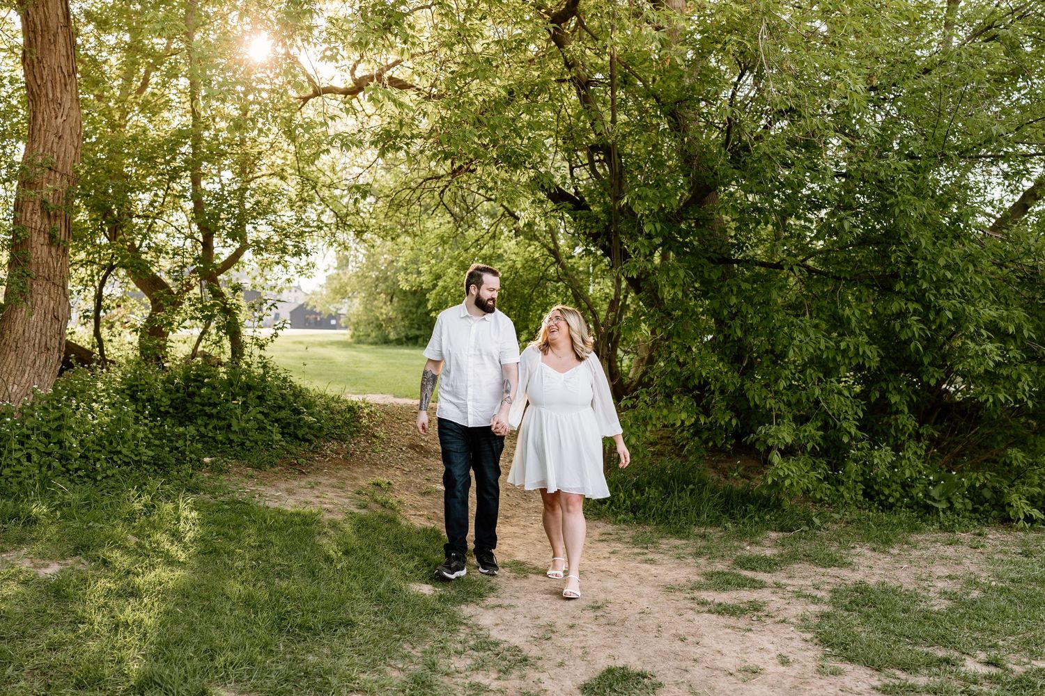Couple holding hands walking on a sunny dirt path surrounded by lush green trees during an outdoor engagement session.