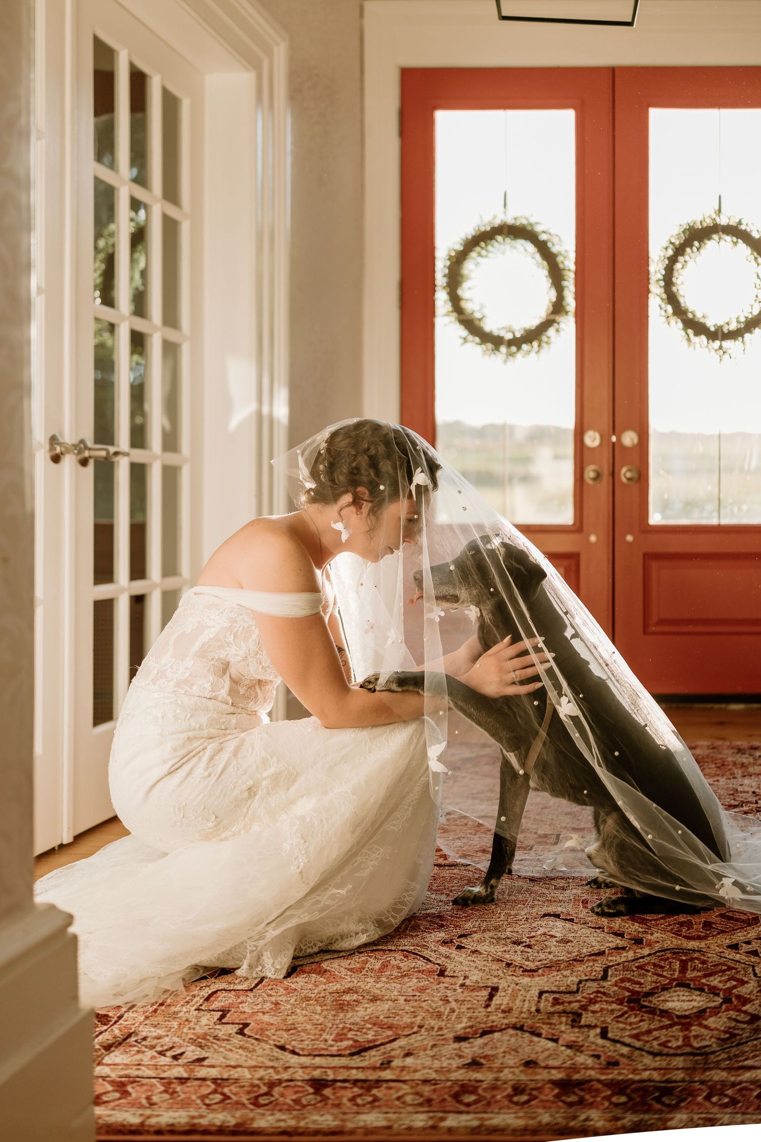 Wedding dress displayed on ornate oriental rug in sunlit hallway with red door.