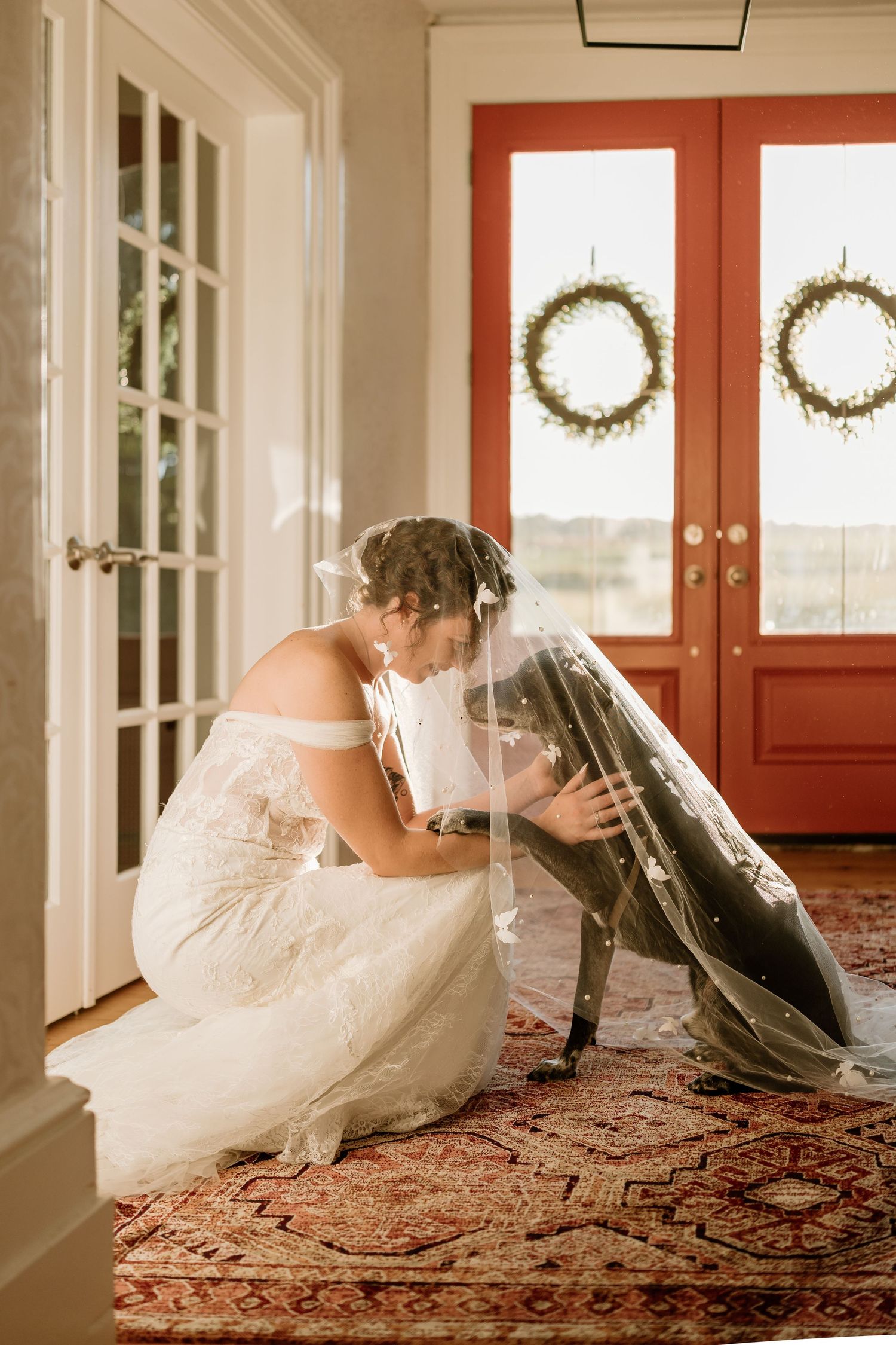 A bride in a white dress kneeling on an ornate rug near red doors with decorative wreaths.