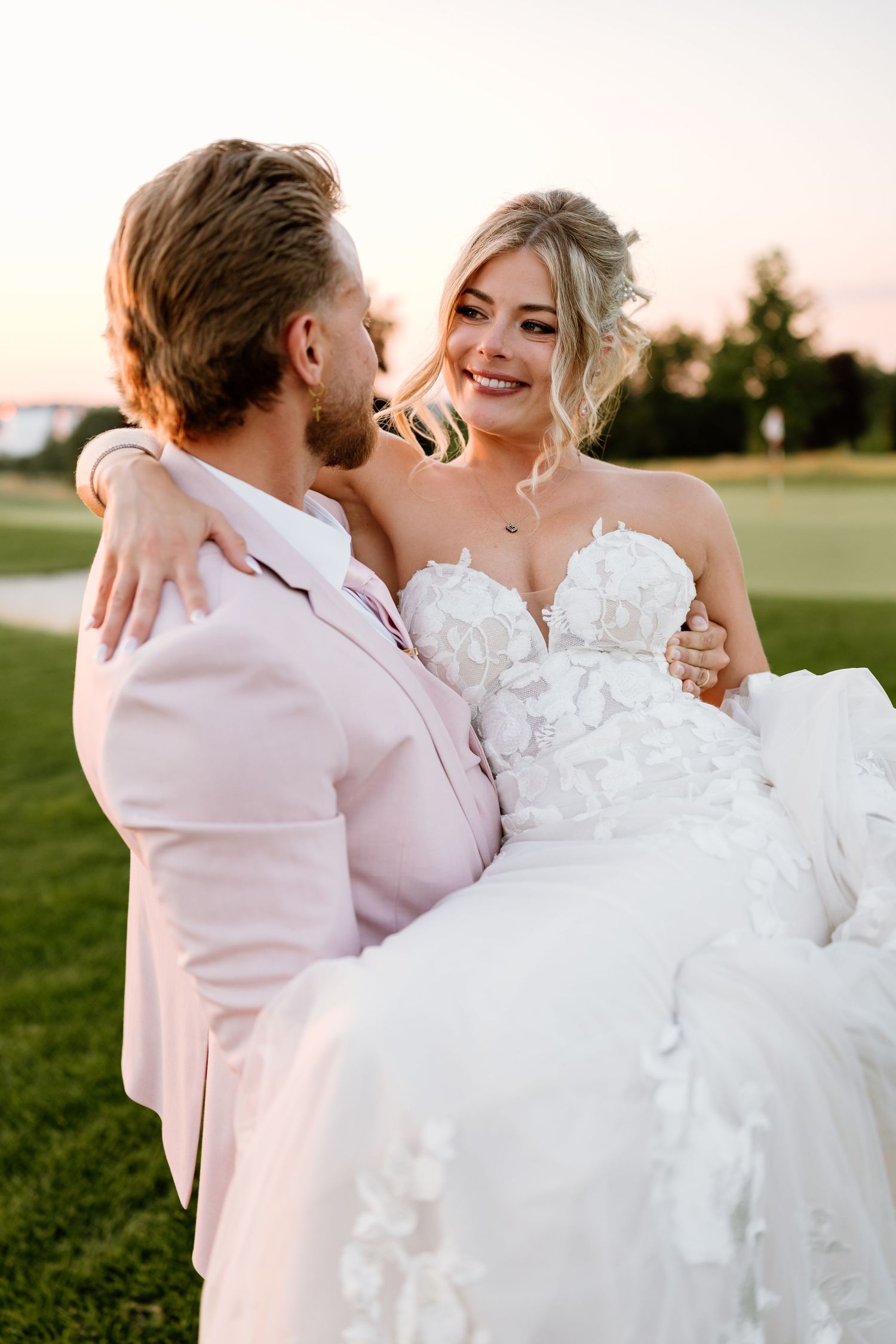 Groom in pink suit carrying smiling bride in white floral gown at sunset.