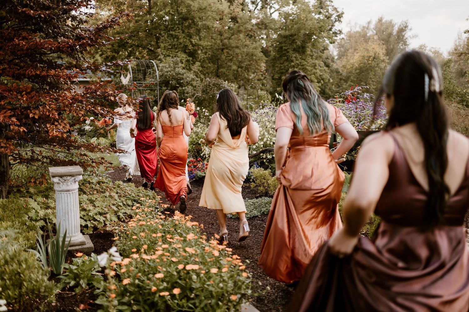 Bridesmaids in flowing dresses walk through a garden path lined with autumn foliage.