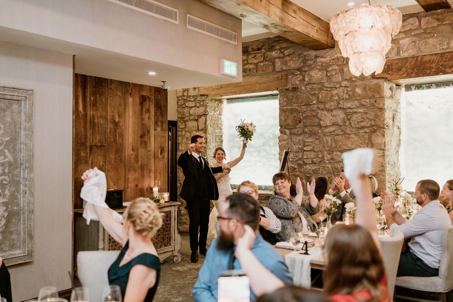 Wedding guests waving napkins during a celebration in a rustic venue with stone walls and wooden beams.