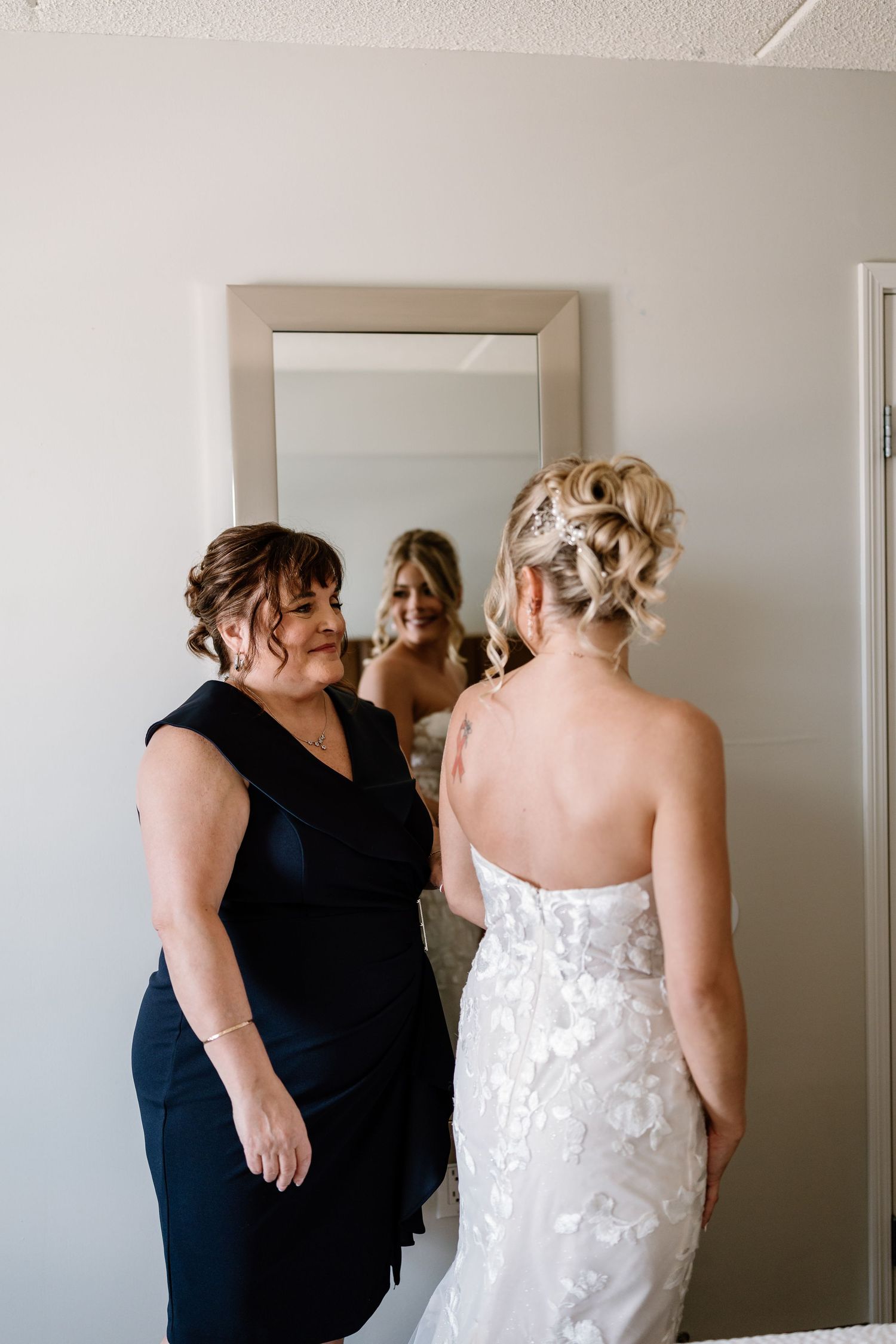 Bride in white lace gown views reflection in mirror with woman in navy dress during wedding preparations.