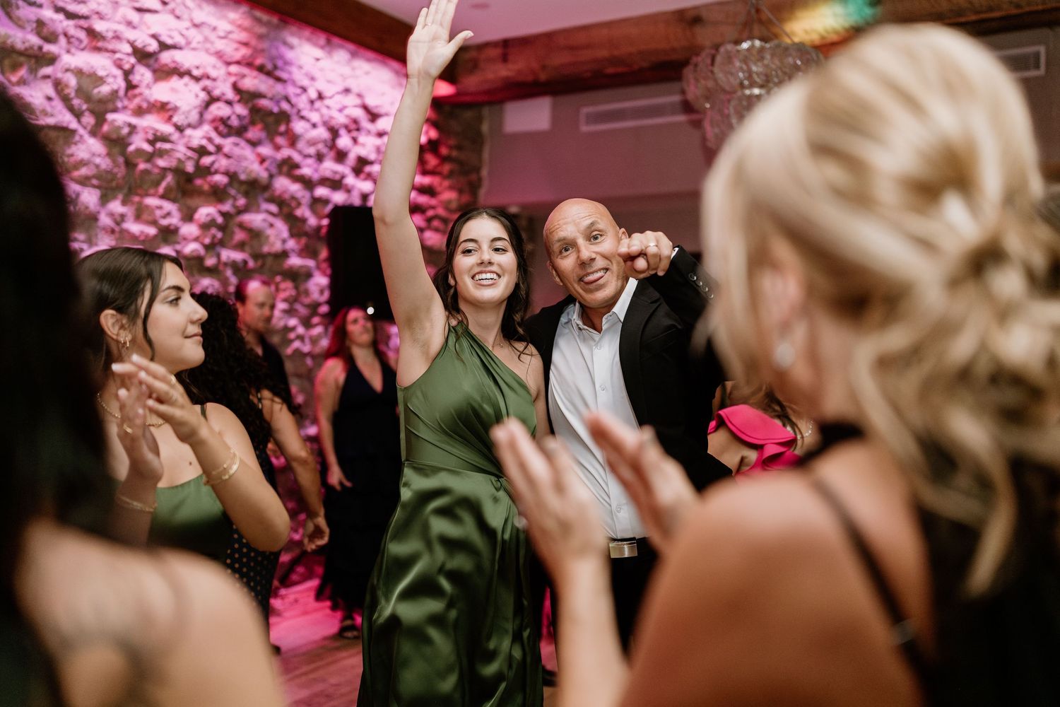 Wedding reception dance party with pink lighting and stone wall backdrop.