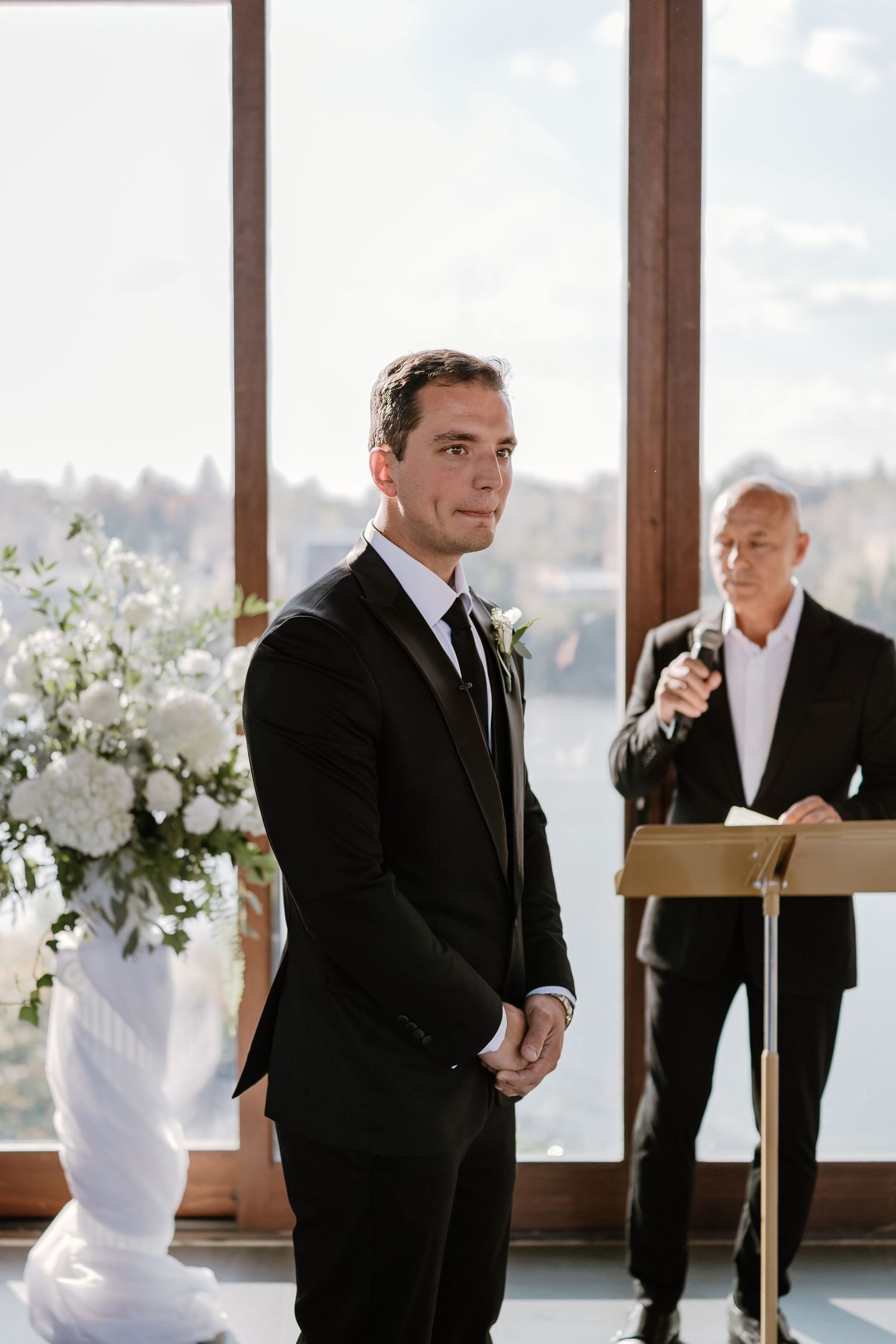 A formal wedding ceremony setup with white floral arrangements and large windows.