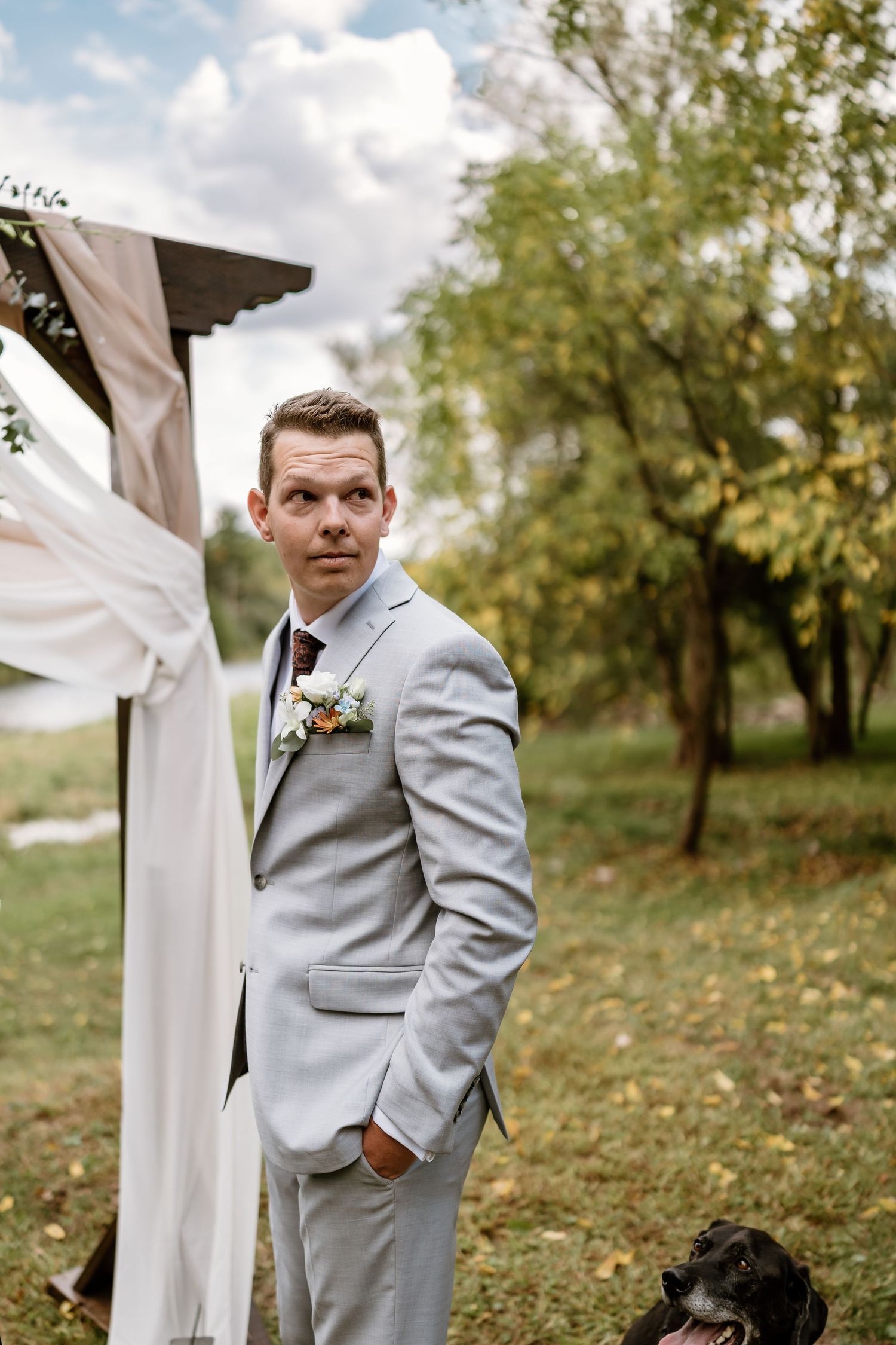 A person in a light gray suit standing by a decorated wedding arch with a black dog nearby.