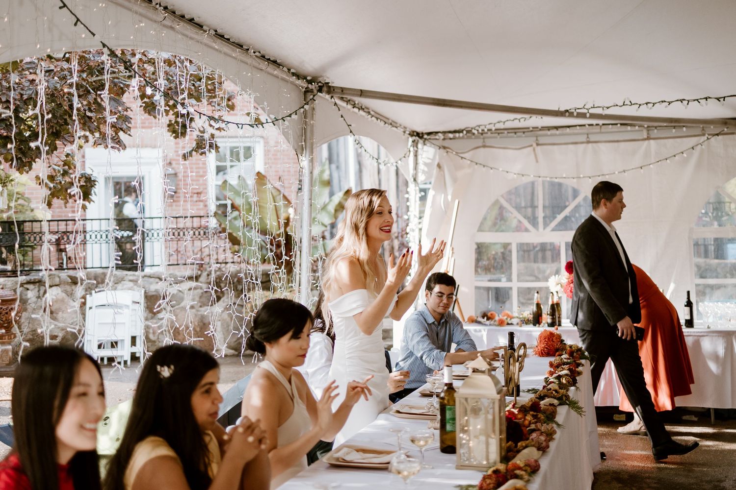 Guests enjoying a celebration in a tent-covered venue with string lights and window views.