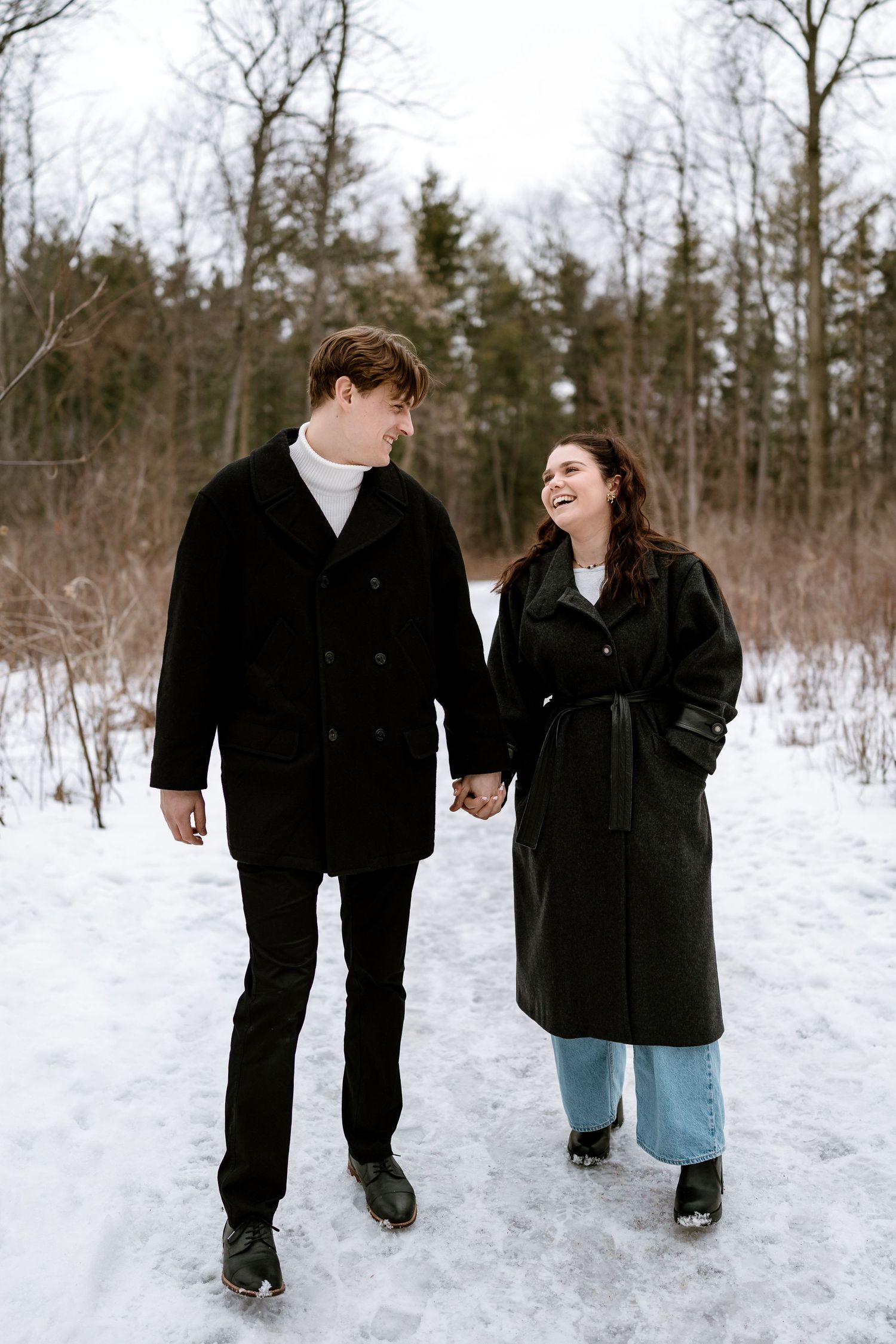 Couple walking hand in hand through snowy winter landscape wearing matching black coats.