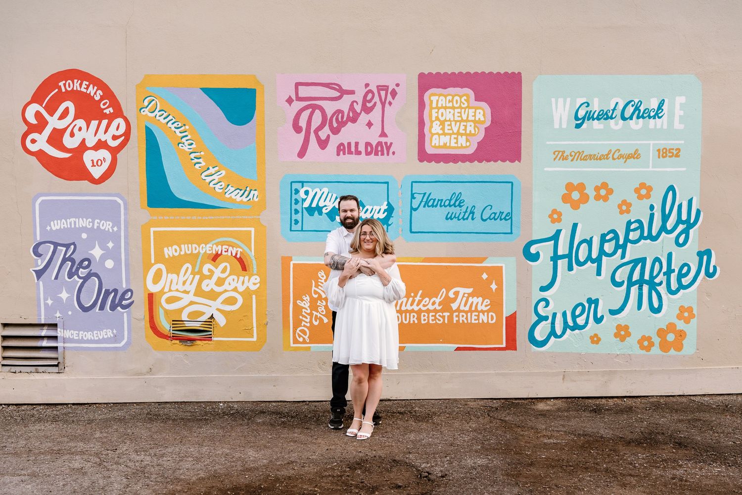 Couple poses in front of a colorful love-themed mural with phrases like Happily Ever After at their engagement shoot.