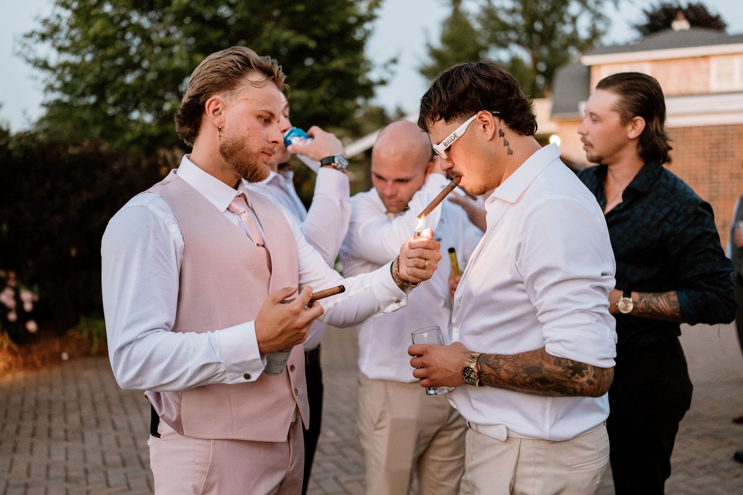 Men in formal attire lighting cigars at an outdoor wedding reception on a sunny evening.