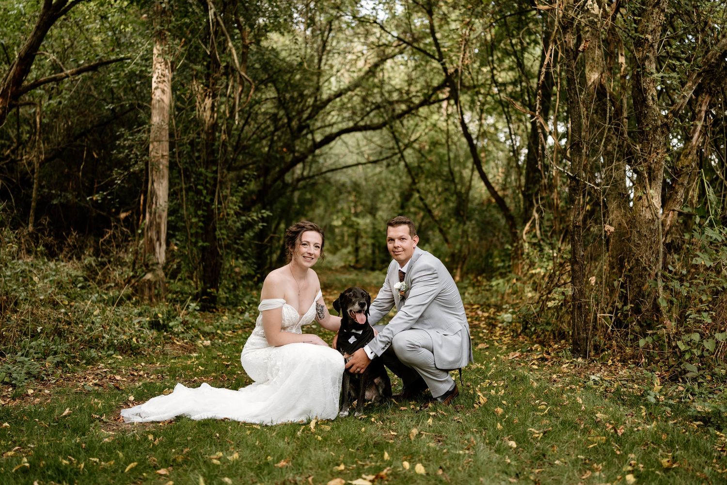 A couple sits with their black dog on grass during their woodland wedding photoshoot.