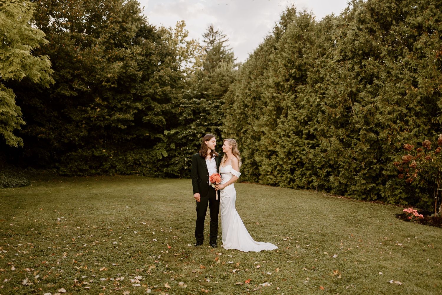 Newlyweds stand together on a lawn with evergreen trees in the background.