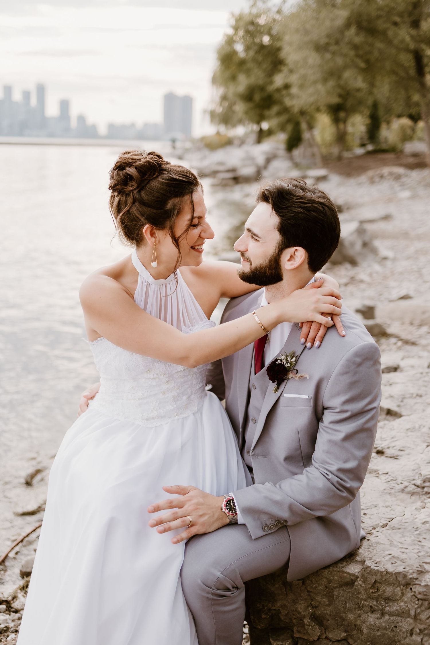 A tender moment between newlyweds on a rocky beach with a city skyline visible.