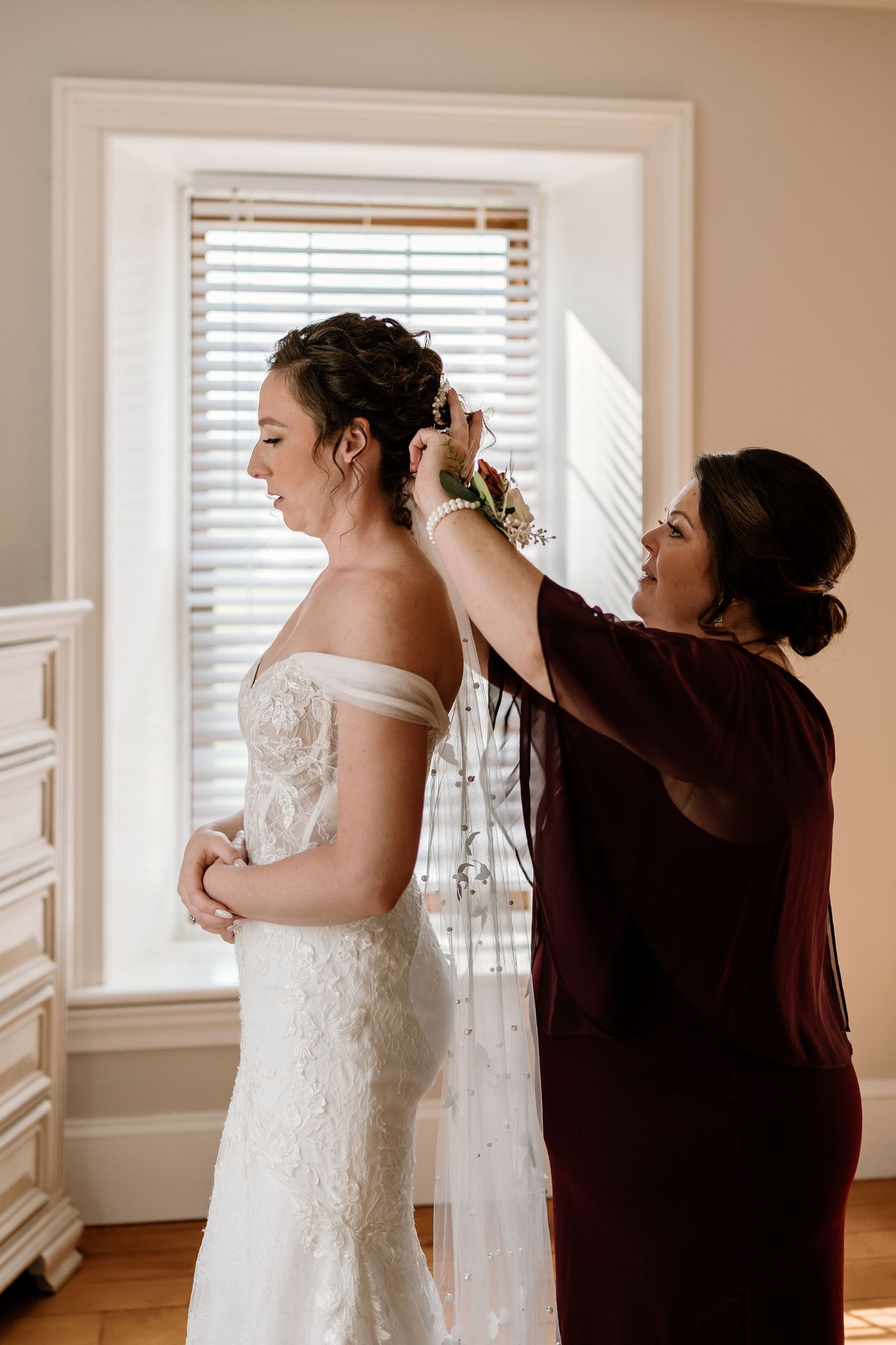 Final adjustments being made to a white lace wedding gown in a sunlit room.