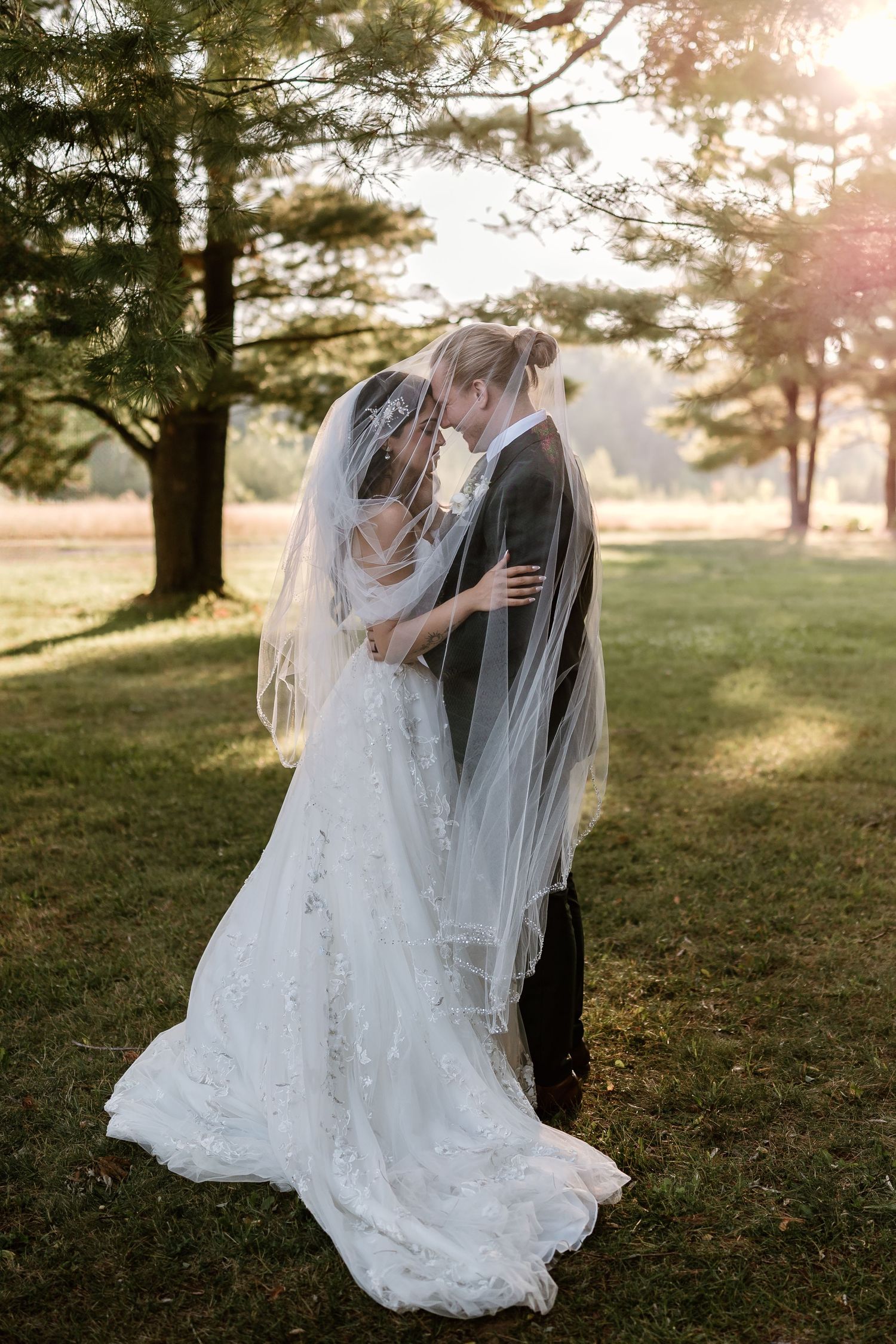Bride and groom share a romantic moment under a flowing veil outdoors, bathed in golden sunlight near lush green trees.