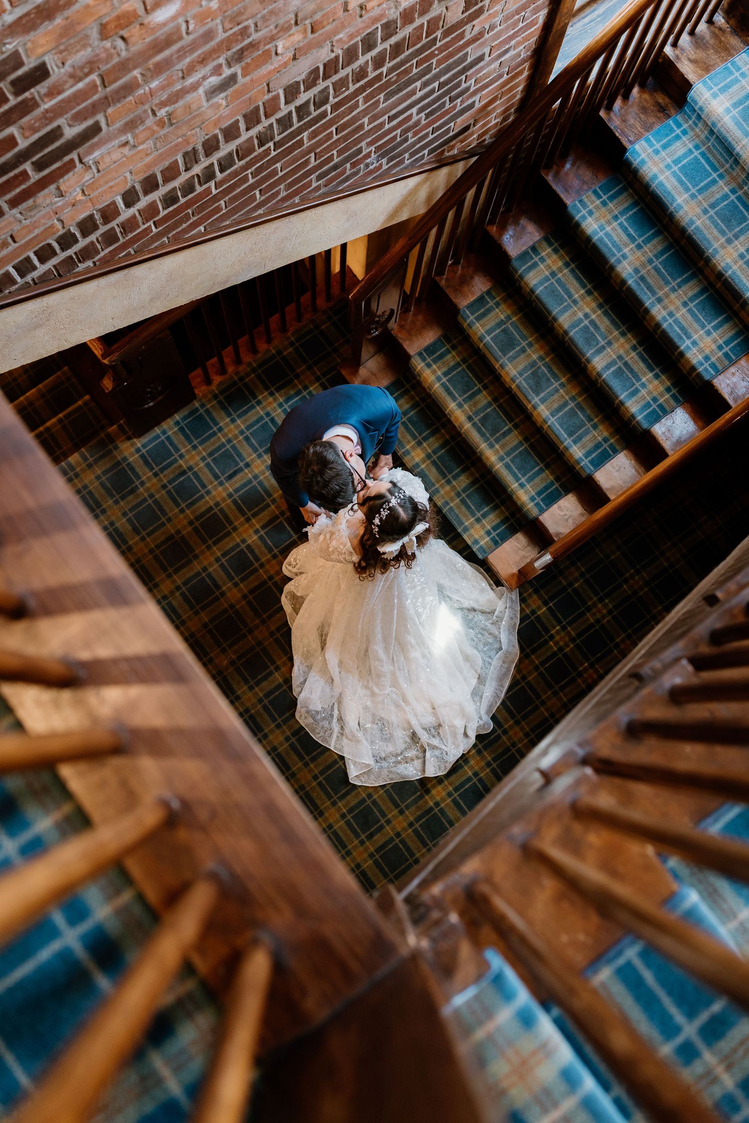 Aerial view of bride and groom embracing on plaid carpet staircase in rustic brick building.