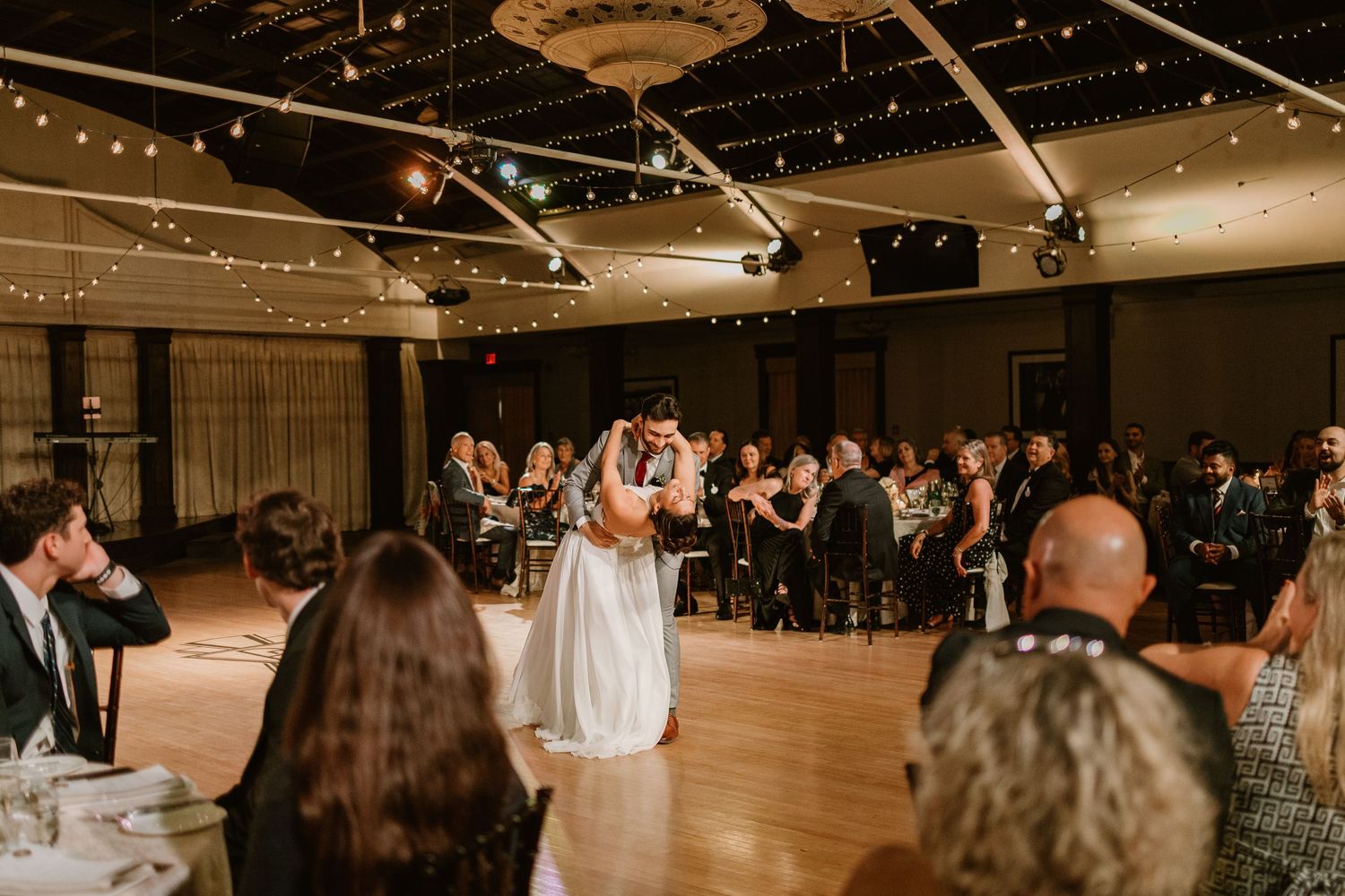 Dimly lit reception hall with string lights and guests dancing on wooden floor.