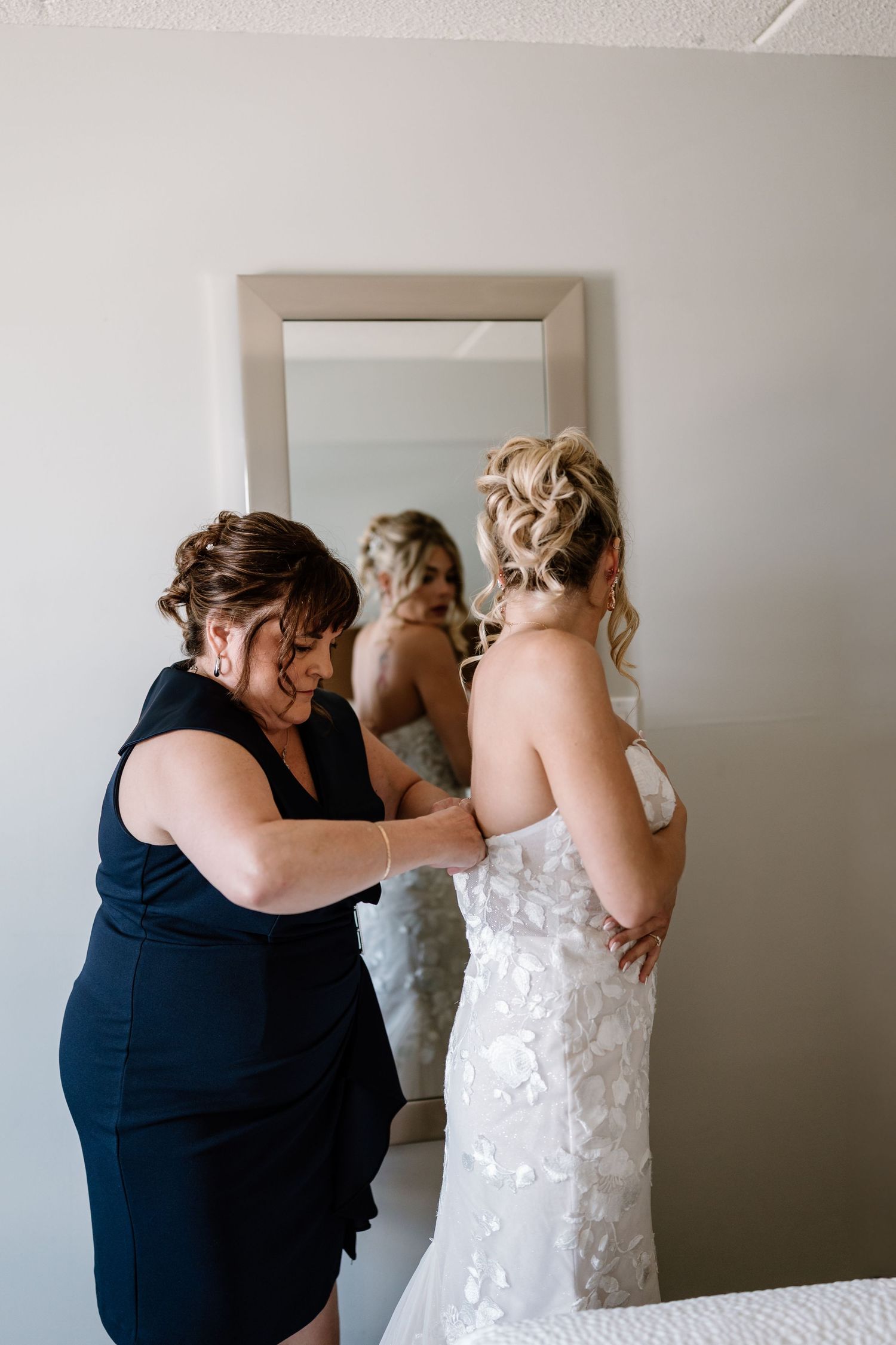 A woman in a navy dress helps a bride in a floral lace gown with her dress in front of a mirror.