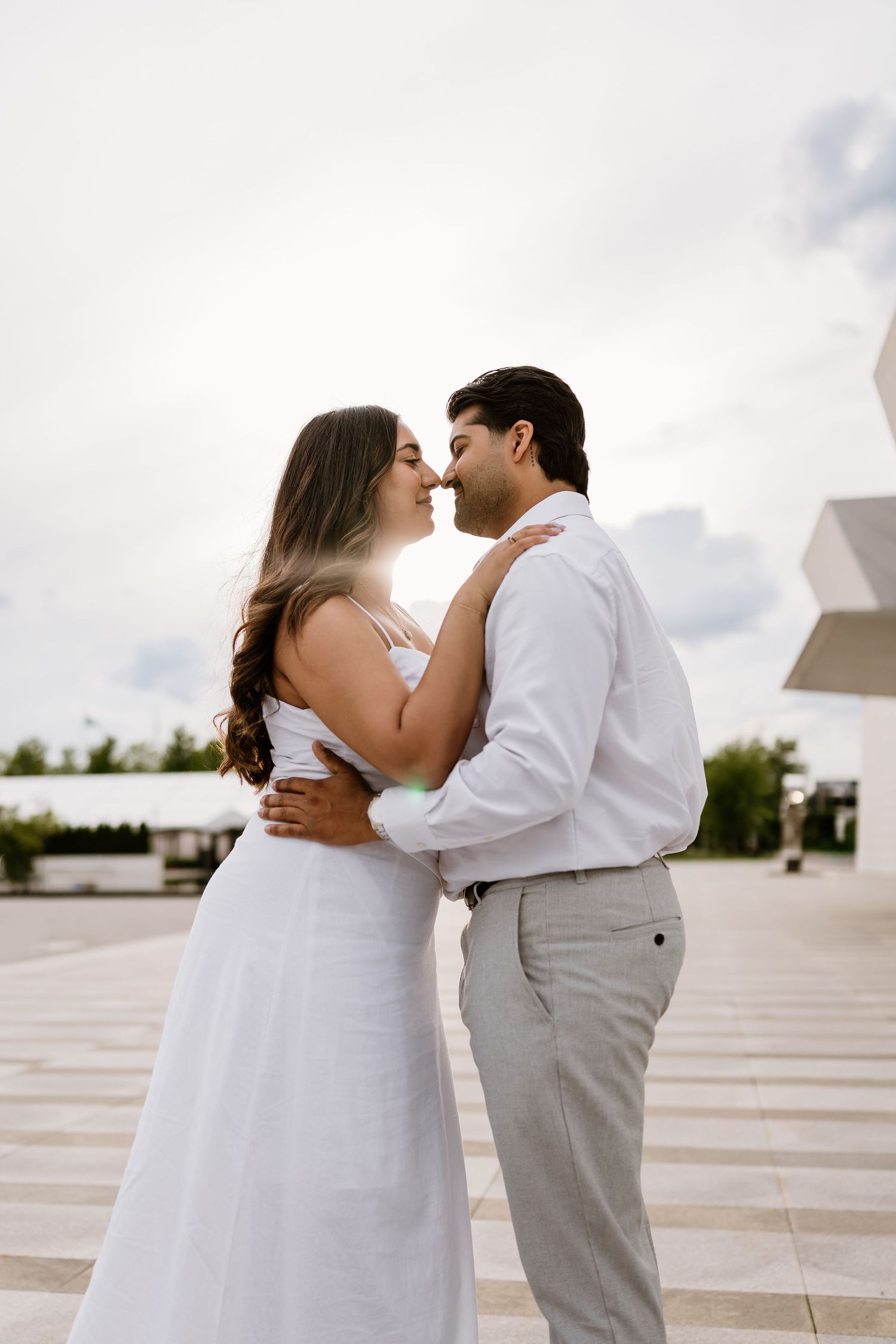 A couple in white wedding attire share a romantic moment, nearly kissing outdoors under a bright cloudy sky.