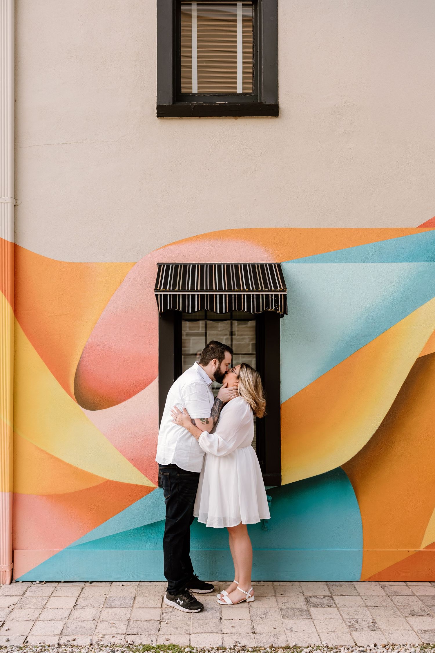 Couple sharing a kiss in front of vibrant colorful mural, both dressed in white, urban outdoor setting.
