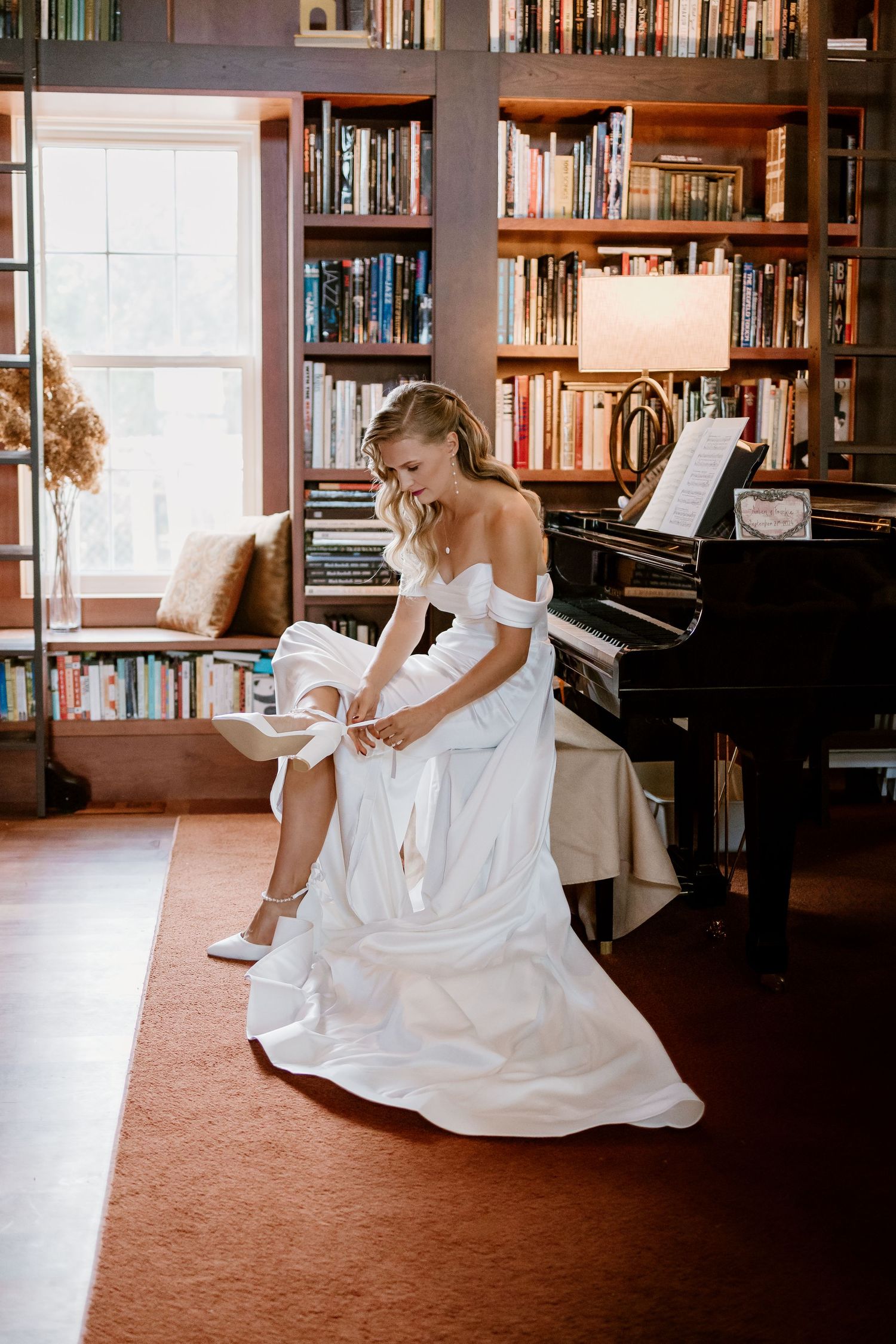 A bride in a white off-shoulder gown adjusts her shoe in a library room with a piano and bookshelves.