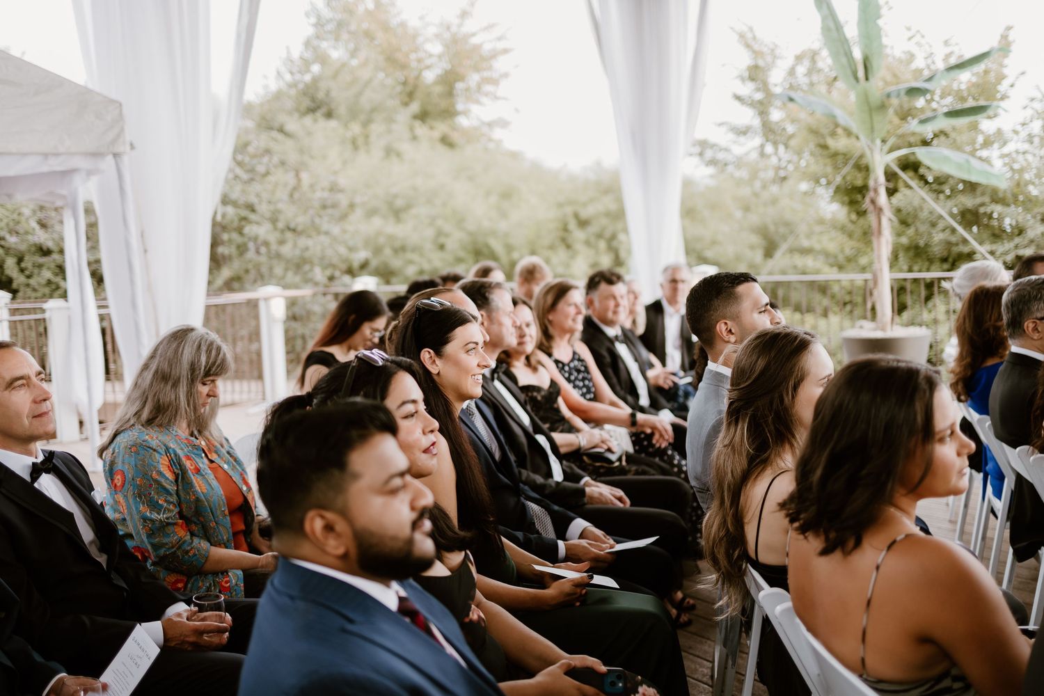 Wedding ceremony guests seated in white chairs under a draped outdoor pavilion.