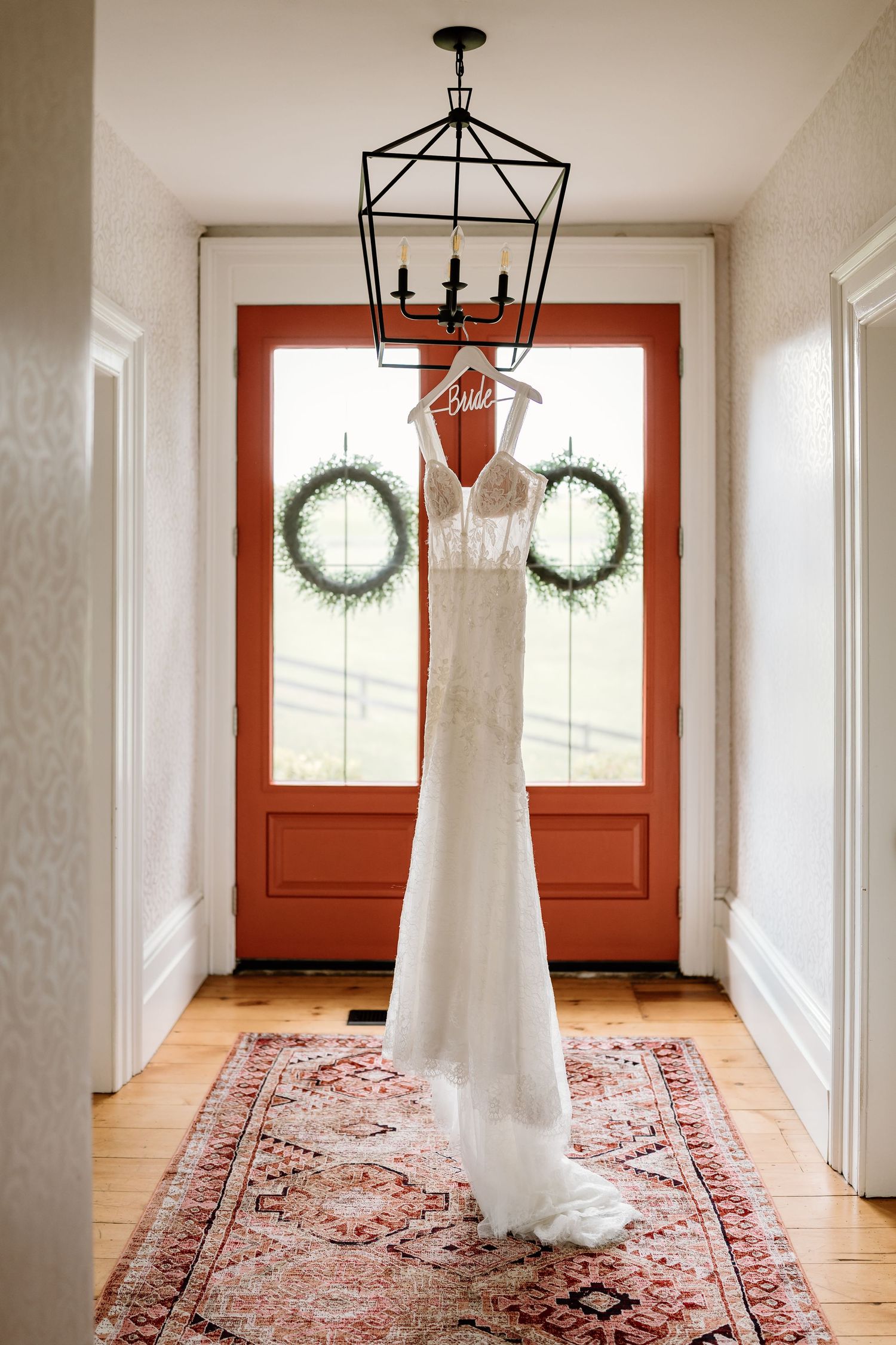 White wedding dress hanging in an entryway with two decorative wreaths on red doors.