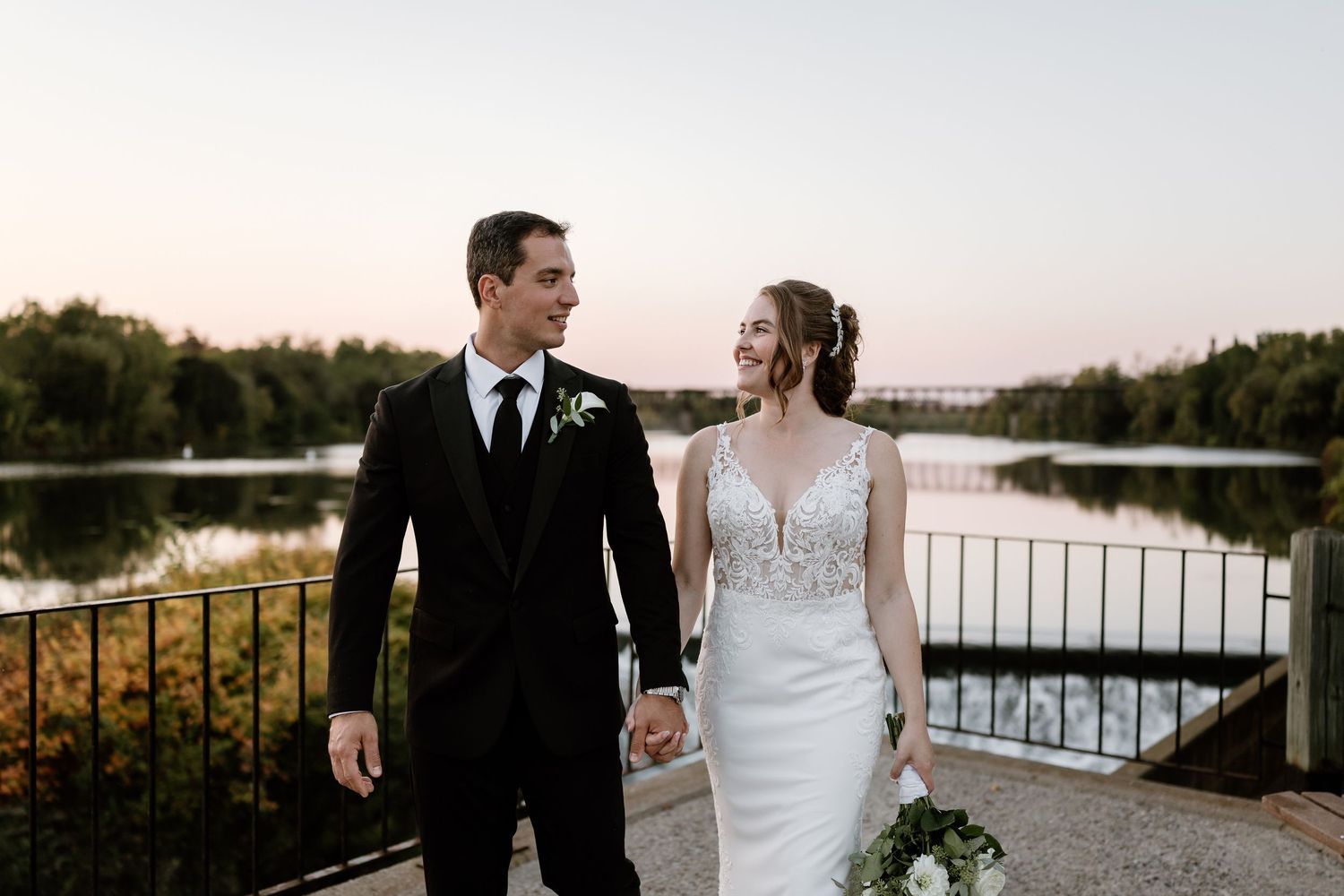 Couple walks along lakeside path during sunset wedding photoshoot.
