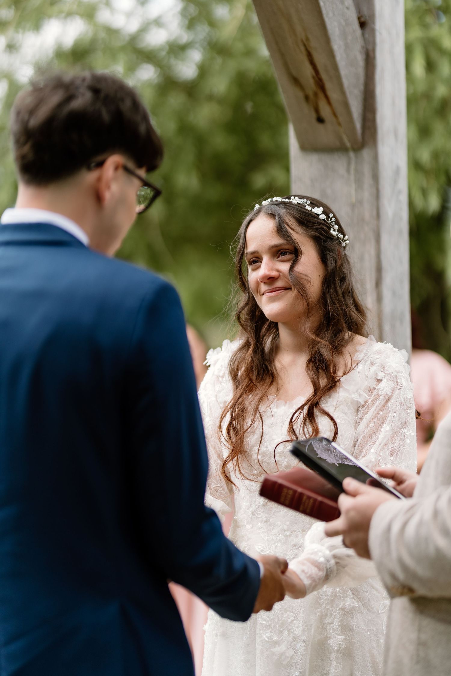 Bride smiling joyfully at groom during outdoor wedding ceremony, wearing white lace dress and floral hair crown.