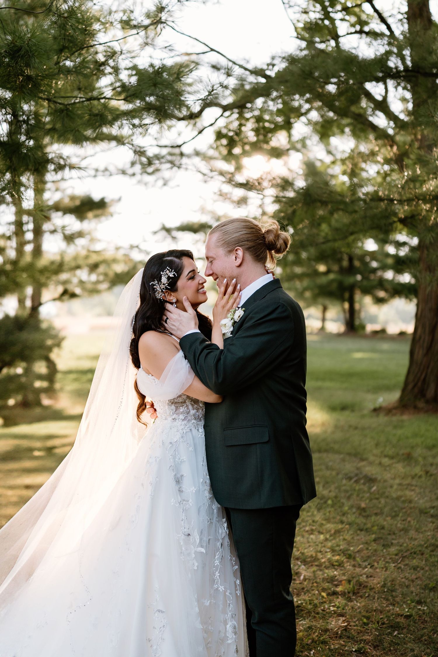 Bride and groom share a romantic moment outdoors, surrounded by lush green trees in golden hour light.