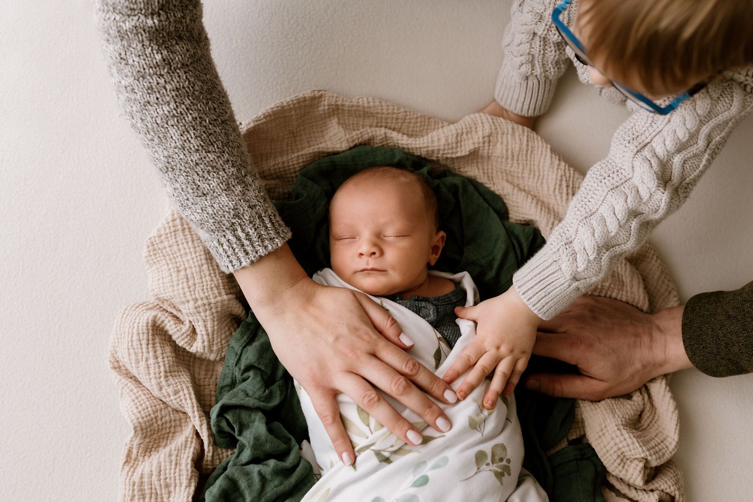 Newborn baby sleeping peacefully wrapped in green blanket while parents and sibling gently place hands on them.
