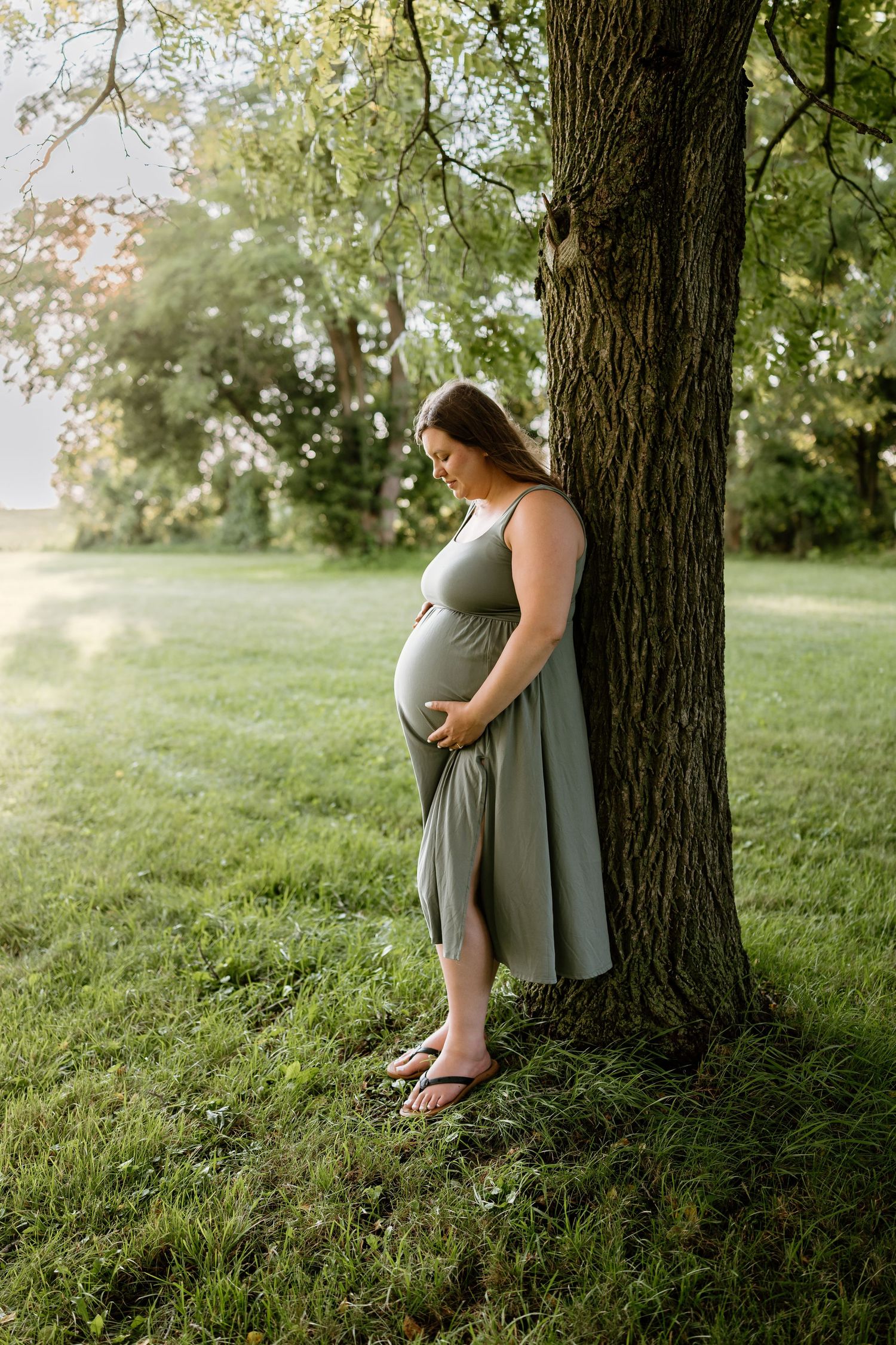 Pregnant woman in sage green dress leaning against a tree in a lush park, cradling her baby bump.