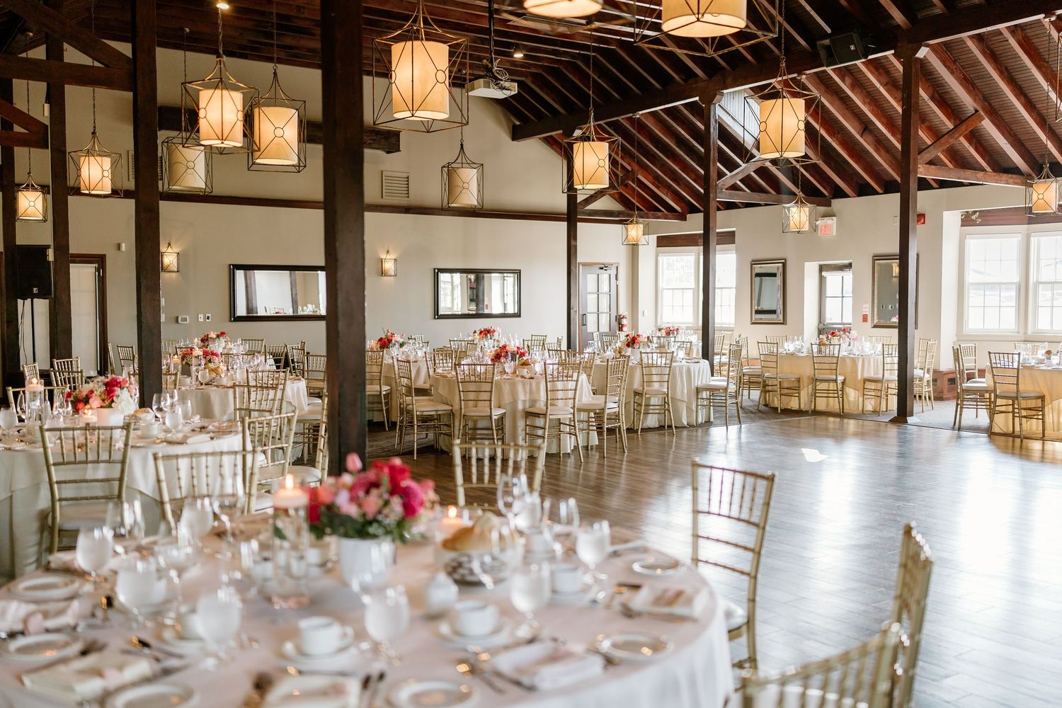 Elegant wedding reception hall with round tables, gold chiavari chairs, pink floral centerpieces, and hanging lanterns.