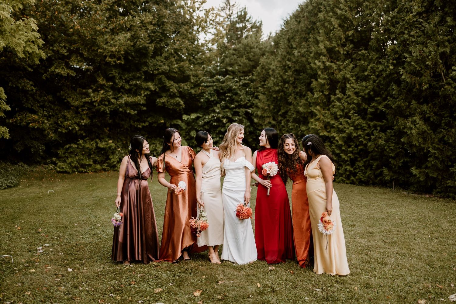 A bridal party in autumn-toned dresses poses together on a green lawn.