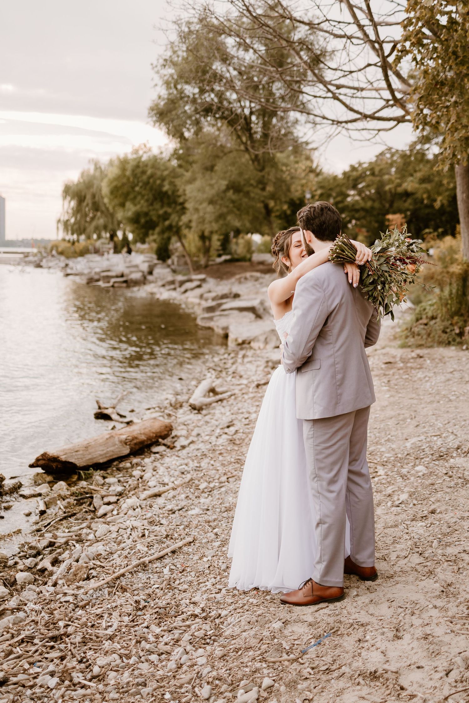A romantic wedding couple embraces on a rocky beach with a city skyline in the distance.