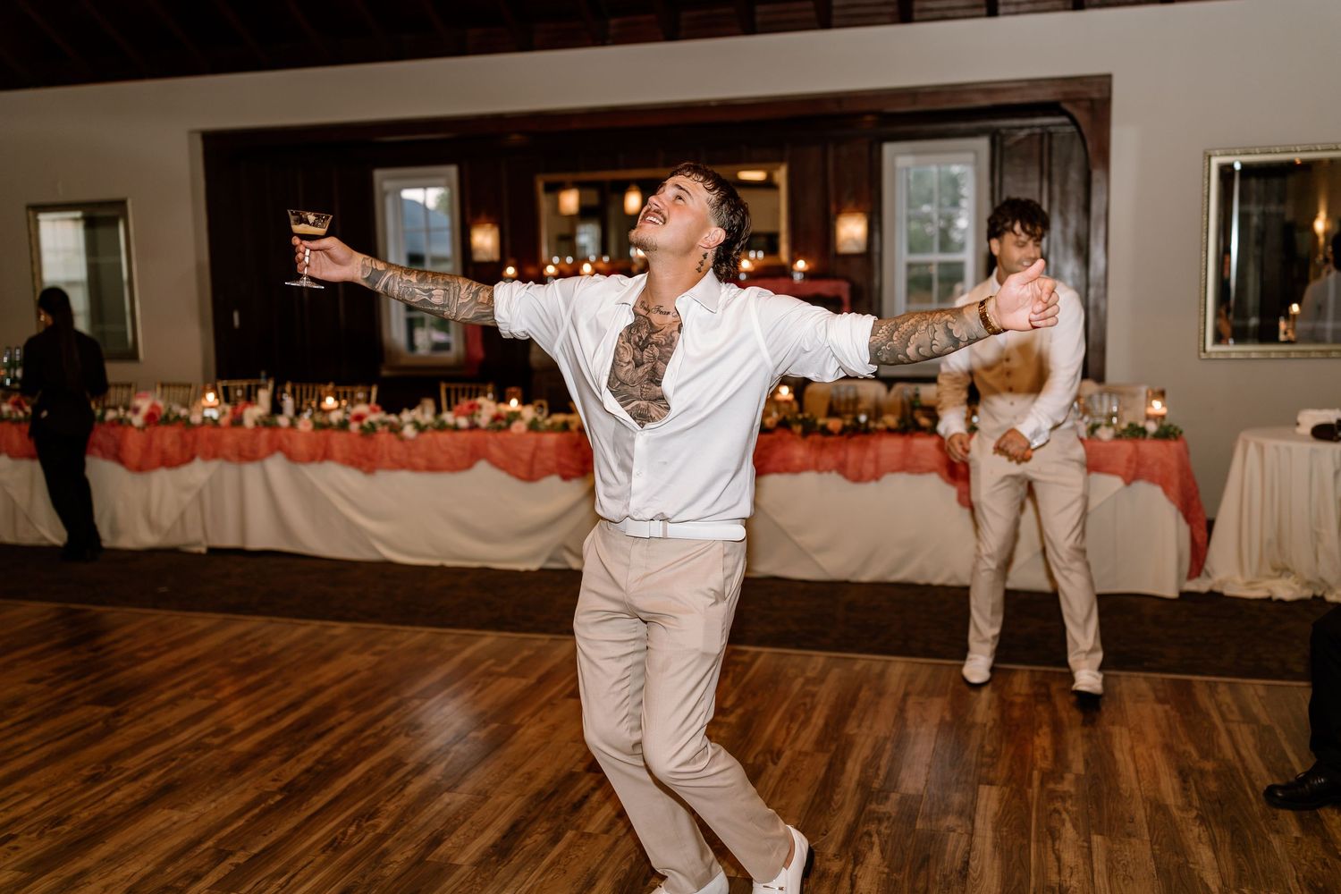 A tattooed man dances joyfully with arms outstretched at a wedding reception on a hardwood dance floor.
