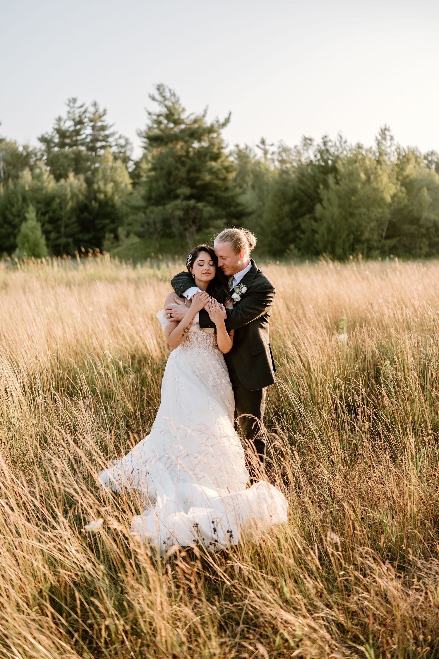Bride and groom embracing in golden meadow, she in lace gown, he in dark suit, with forest backdrop.