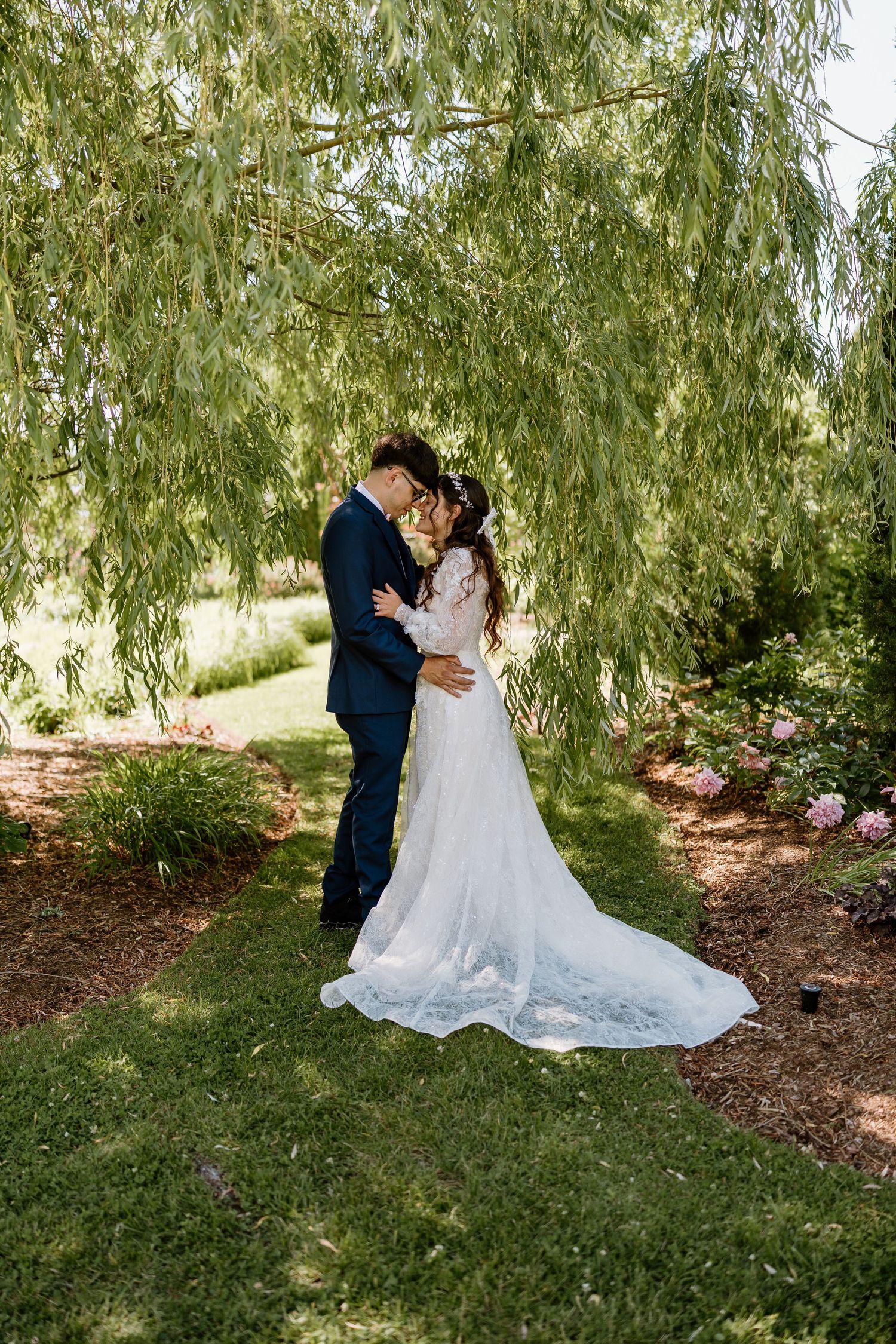 Bride in lace gown and groom in navy suit embrace tenderly under a weeping willow tree in a garden.