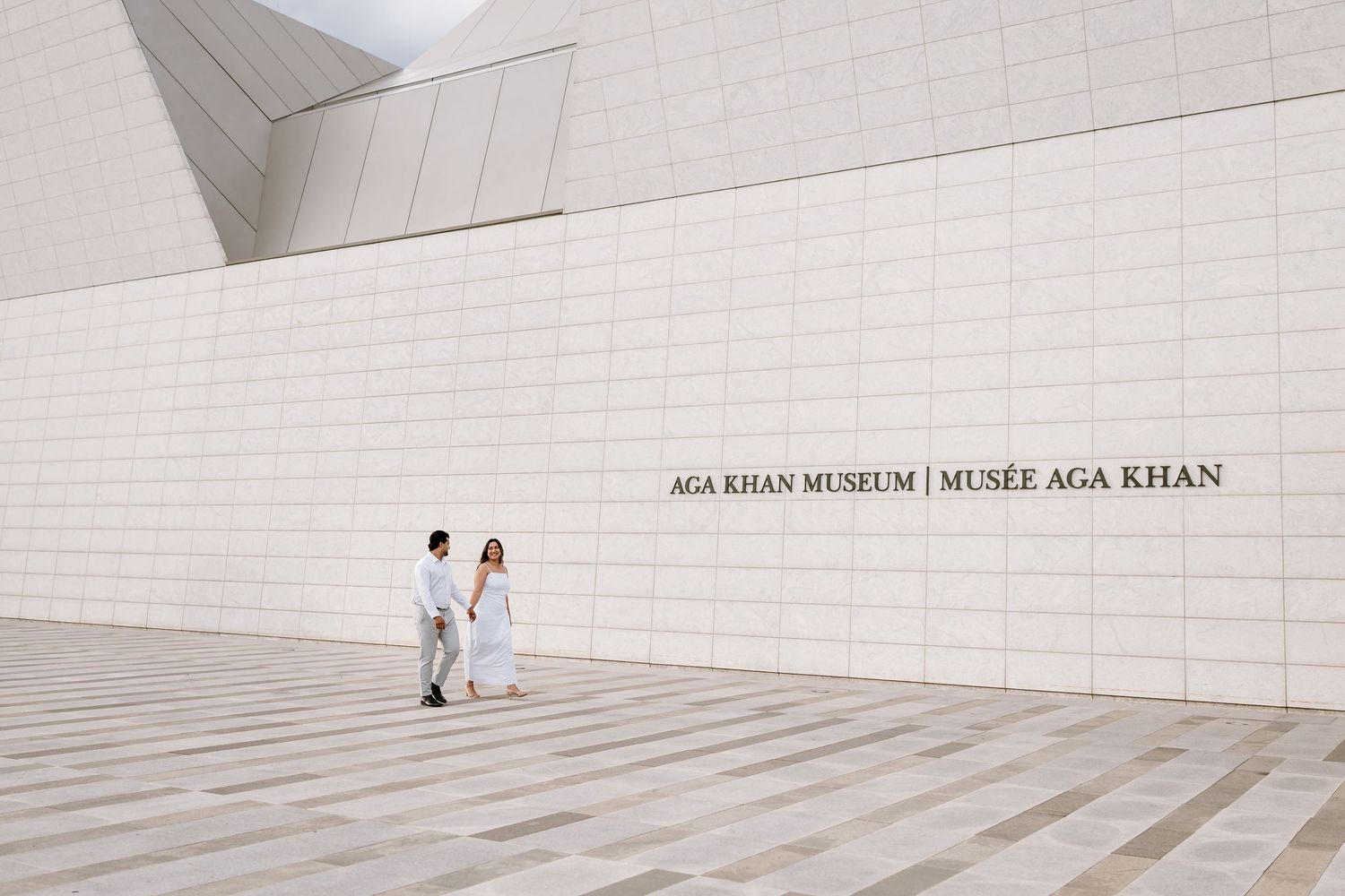 Couple in white attire walking outside the Aga Khan Museum in Toronto on a sunny day.