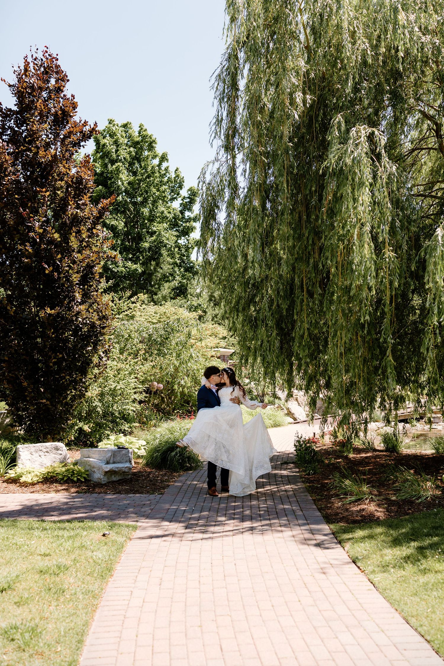 Groom lifts bride in white gown on brick path surrounded by lush green willow and trees on sunny wedding day.