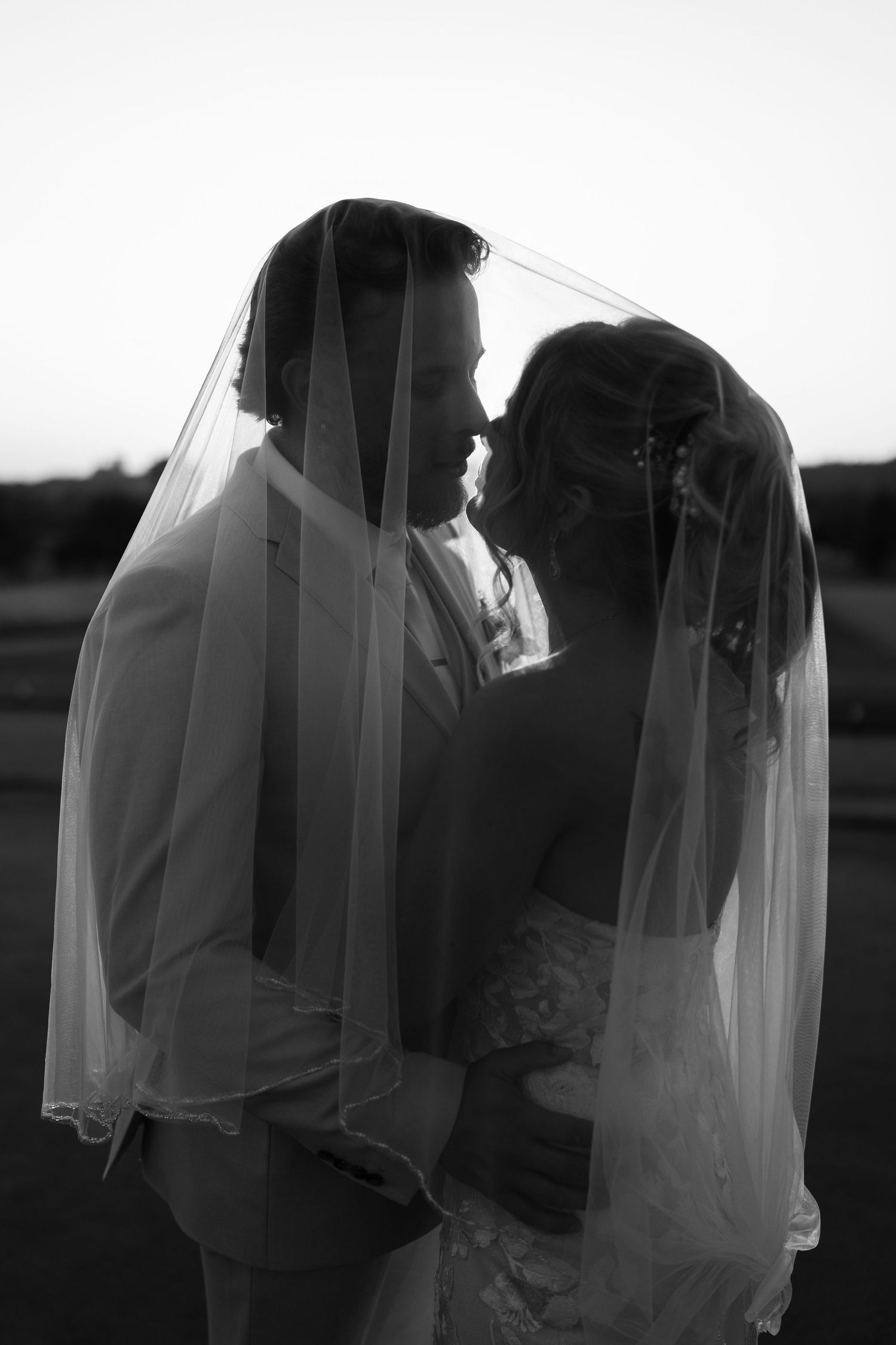 Black and white photo of bride and groom sharing an intimate moment under a wedding veil at sunset.