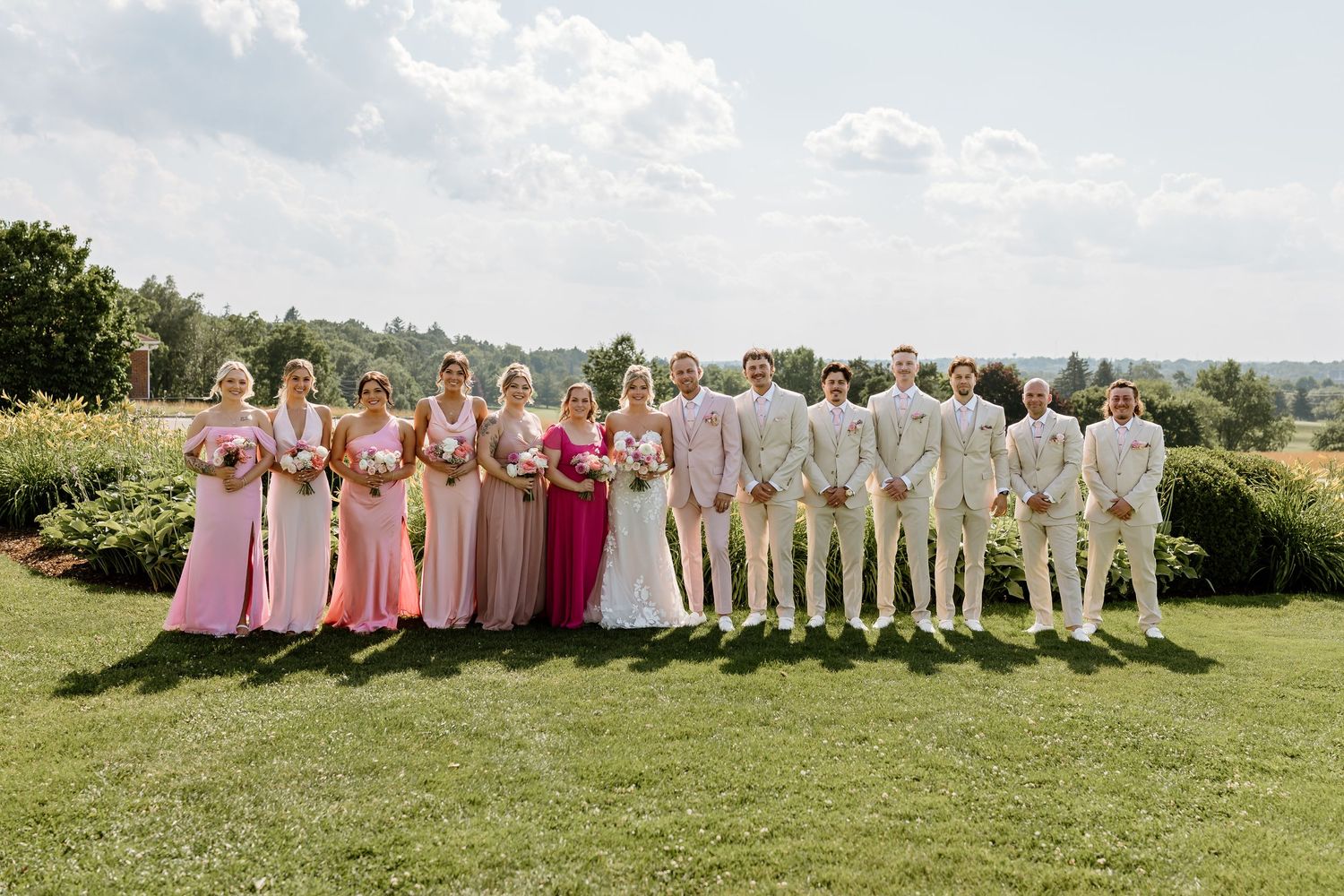 Wedding party poses outdoors on green lawn, bridesmaids in pink gowns and groomsmen in tan suits on a sunny day.