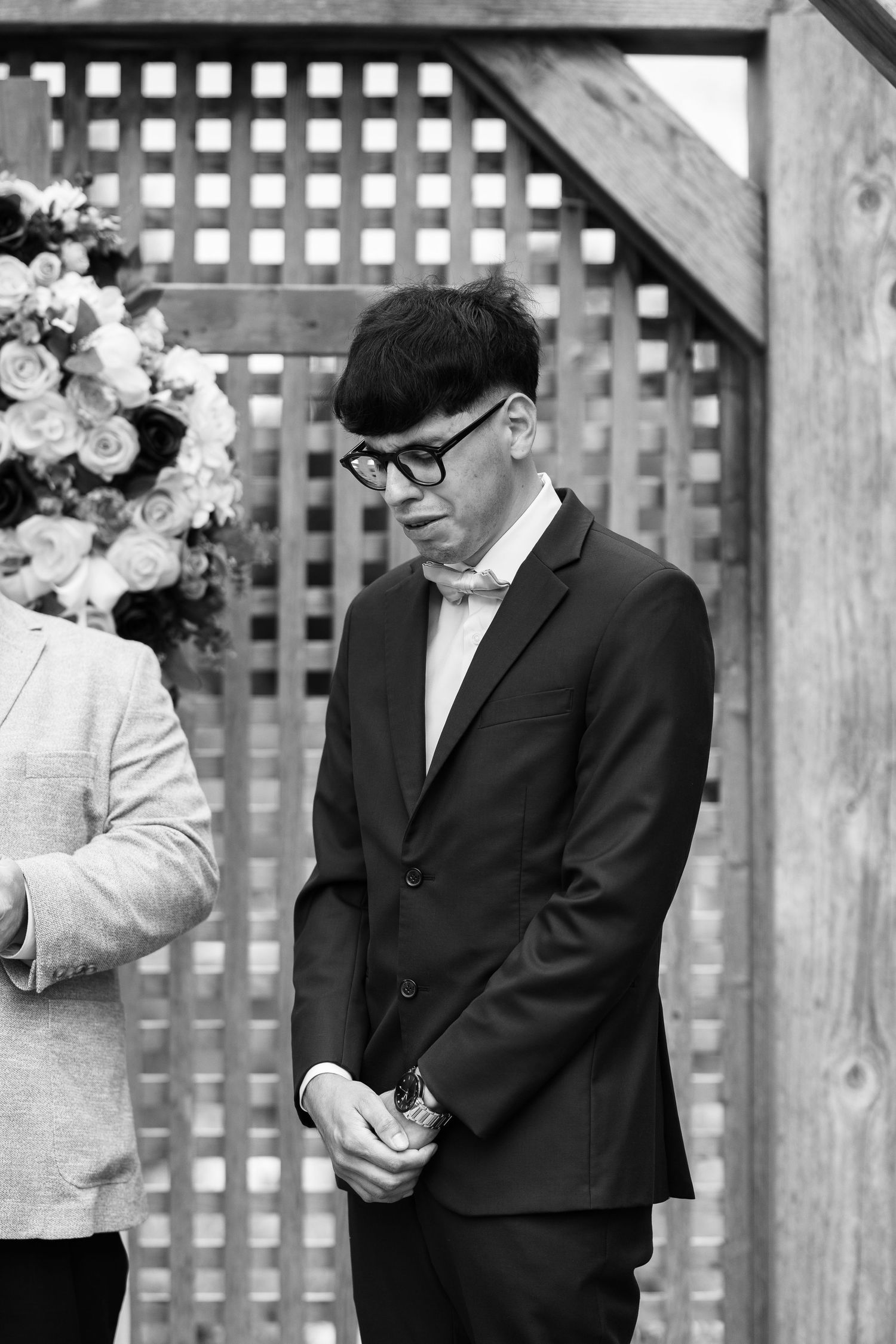 Black and white photo of a man in a dark suit and glasses standing at an outdoor wedding ceremony.