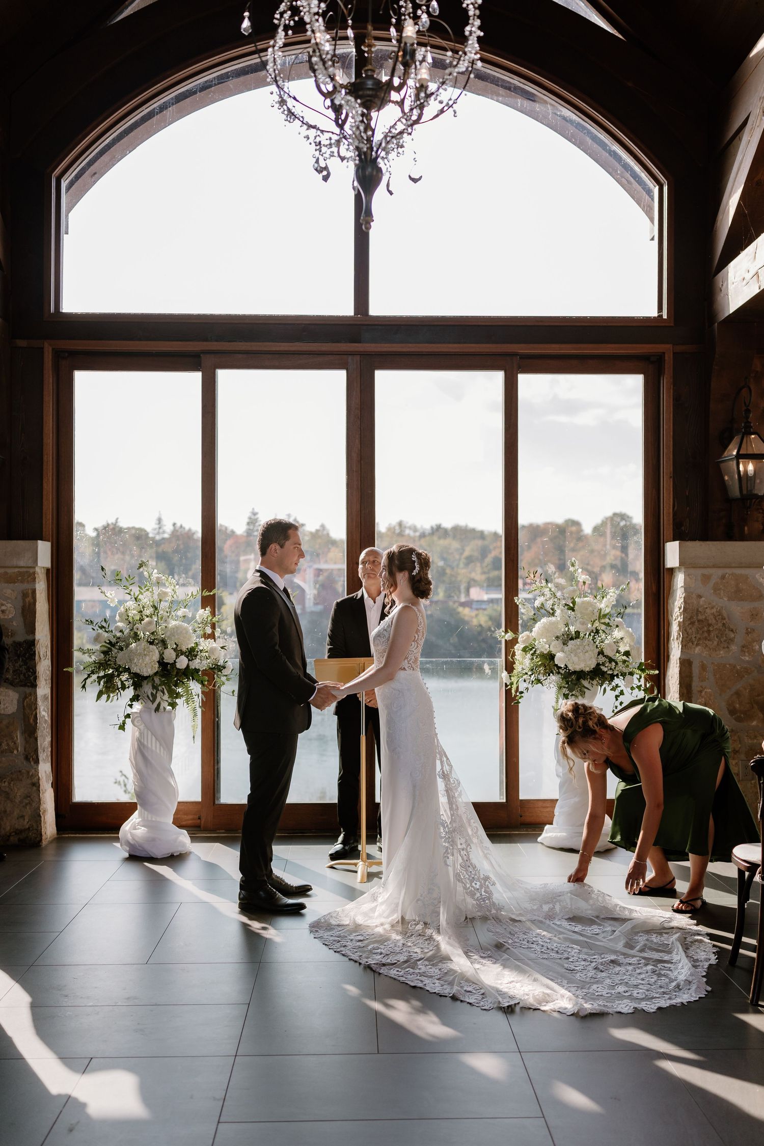 A wedding ceremony in a sunlit room with large windows and elegant floral arrangements.