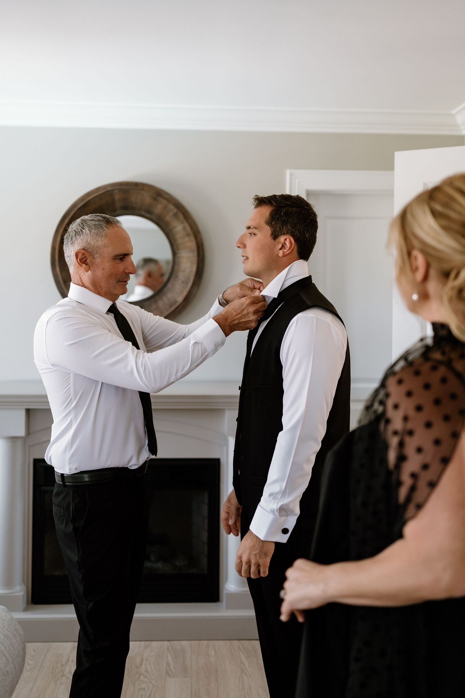 Two people helping with formal attire adjustments in front of a round mirror and fireplace.
