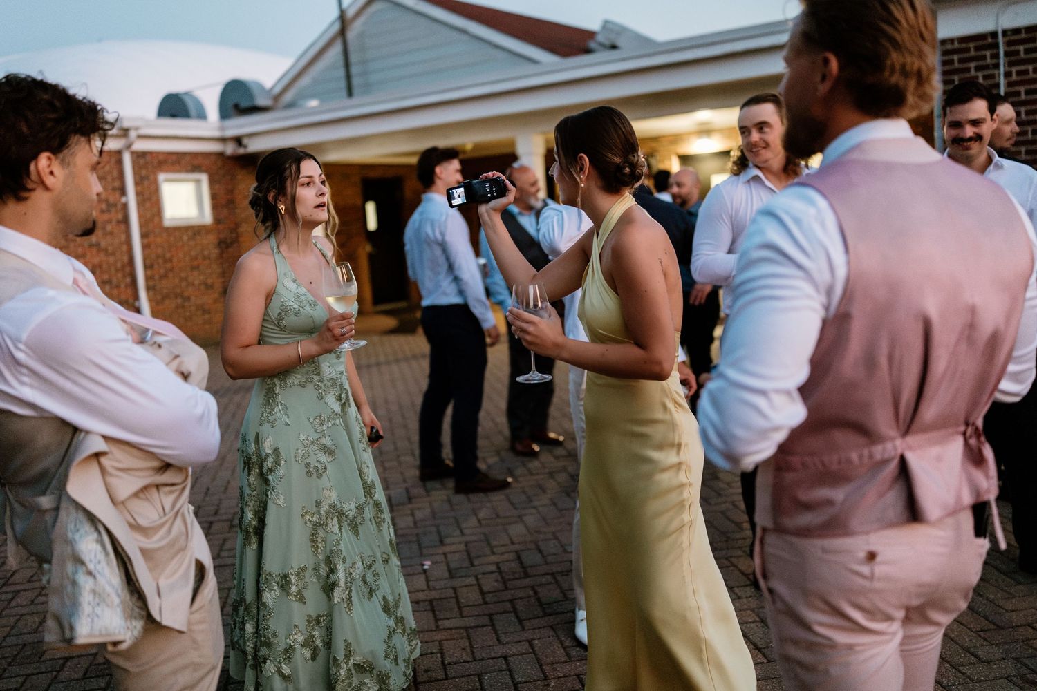 Guests in formal attire socializing outdoors at a wedding reception, wearing floral and pastel dresses and suits.