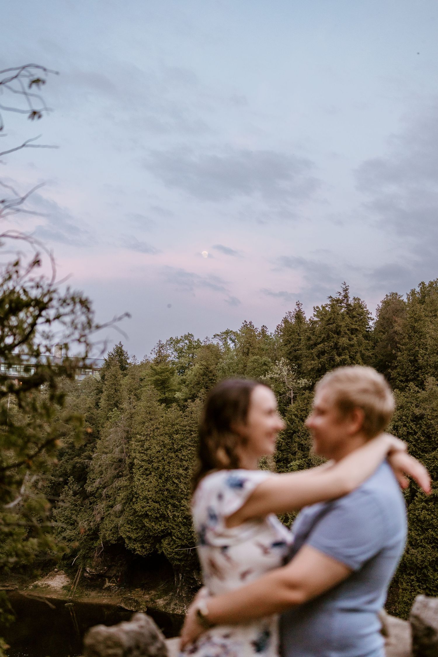 A romantic couple embraces against a forested backdrop with pink sunset skies.
