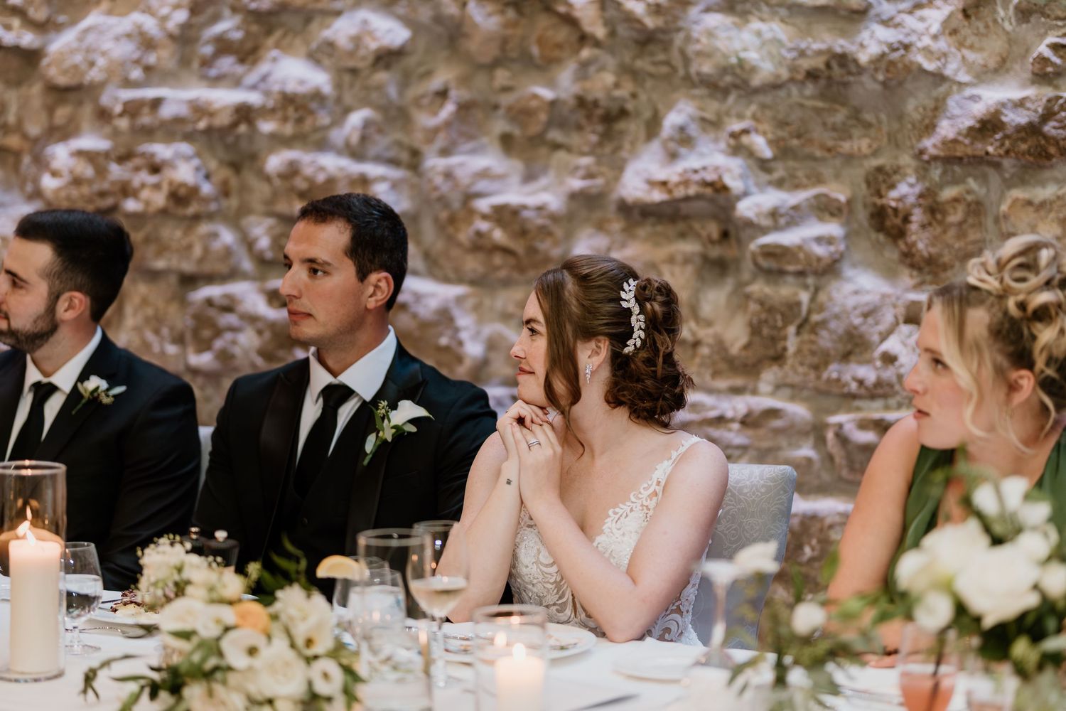 Wedding party seated at head table during reception with stone wall backdrop.